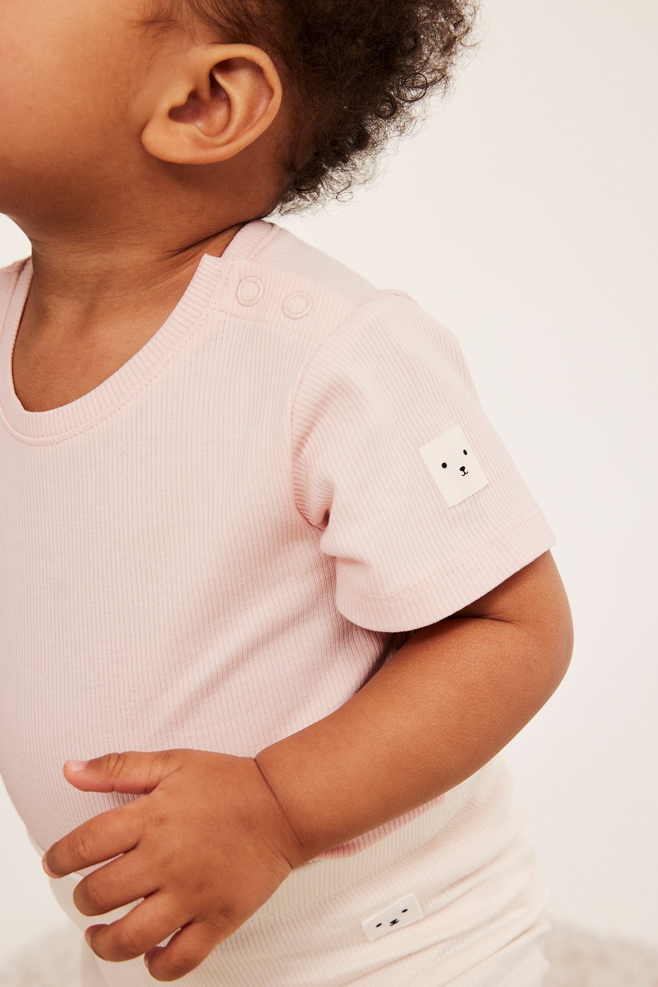 Lähikuva vauvasta yllään vaaleanpunainen ribattu lyhythihainen body olkapään napeilla ja karhutunnuksella.