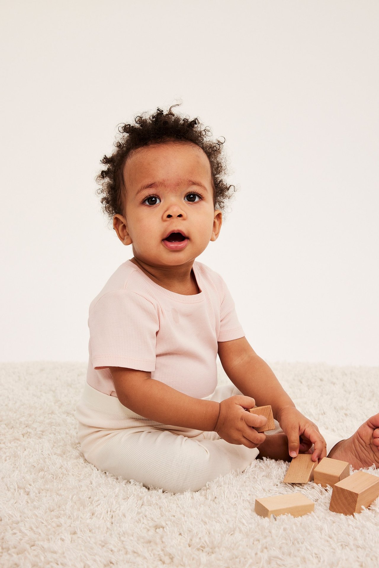Edestä, vauva yllään vaaleanpunainen ribattu lyhythihainen body ja vaaleat housut, leikkii palikoilla.