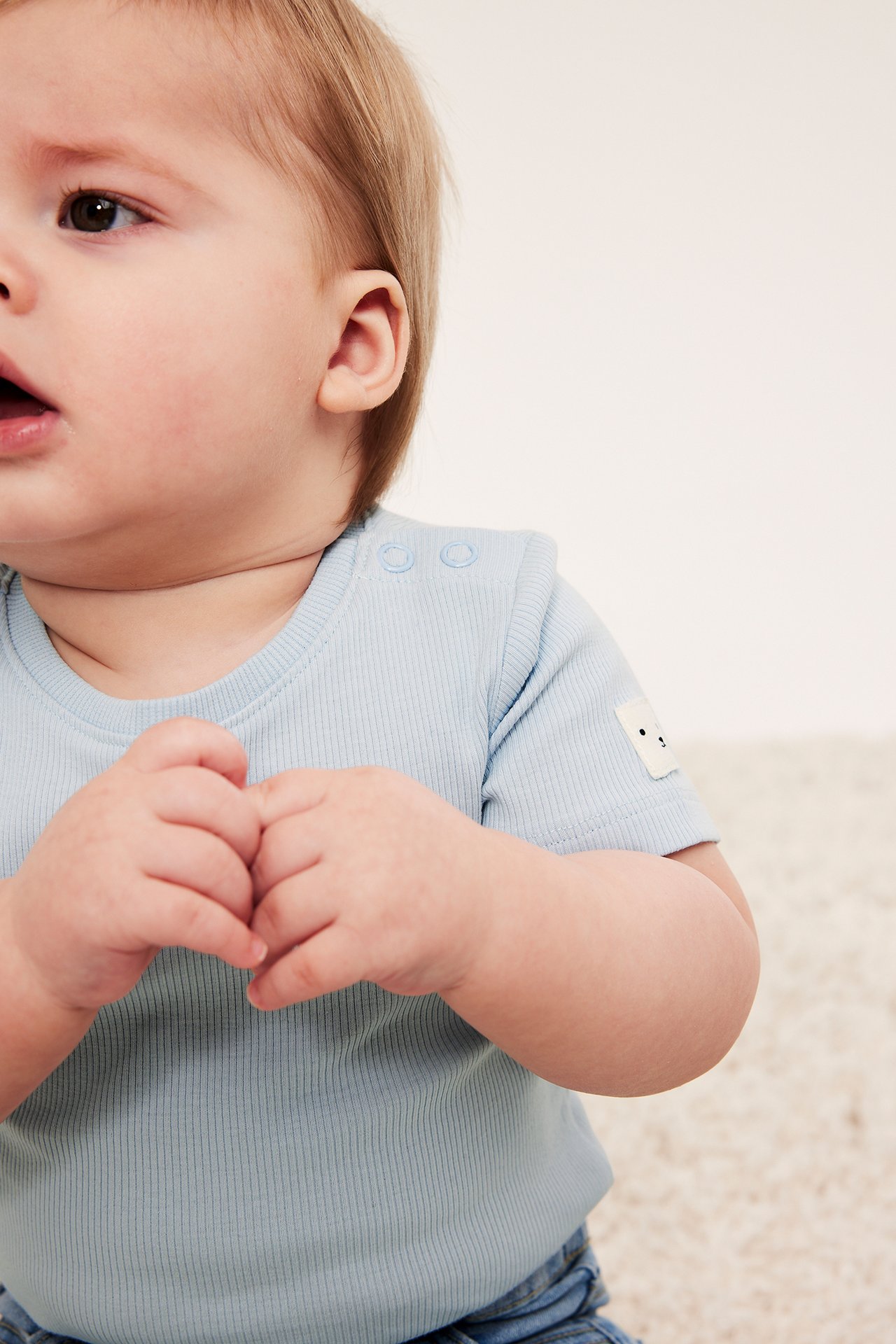Lähikuva sivukuva vauvasta yllään vaaleansininen ribattu lyhythihainen body olkapääneppareilla ja pienellä kasvomerkillä.