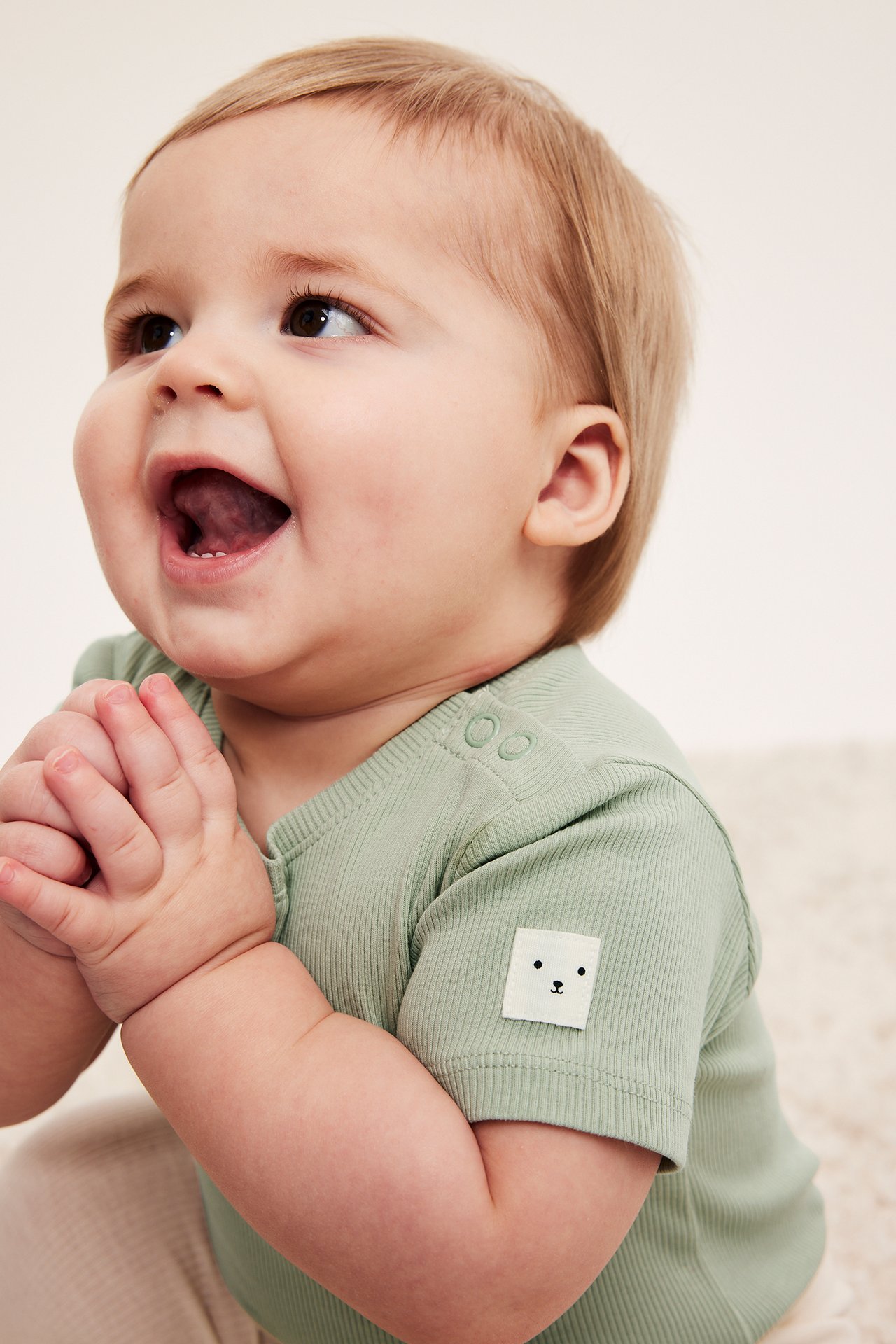 Uśmiechnięte dziecko w jasnozielonym prążkowanym body z krótkim rękawem, napami na ramieniu i naszywką misia.