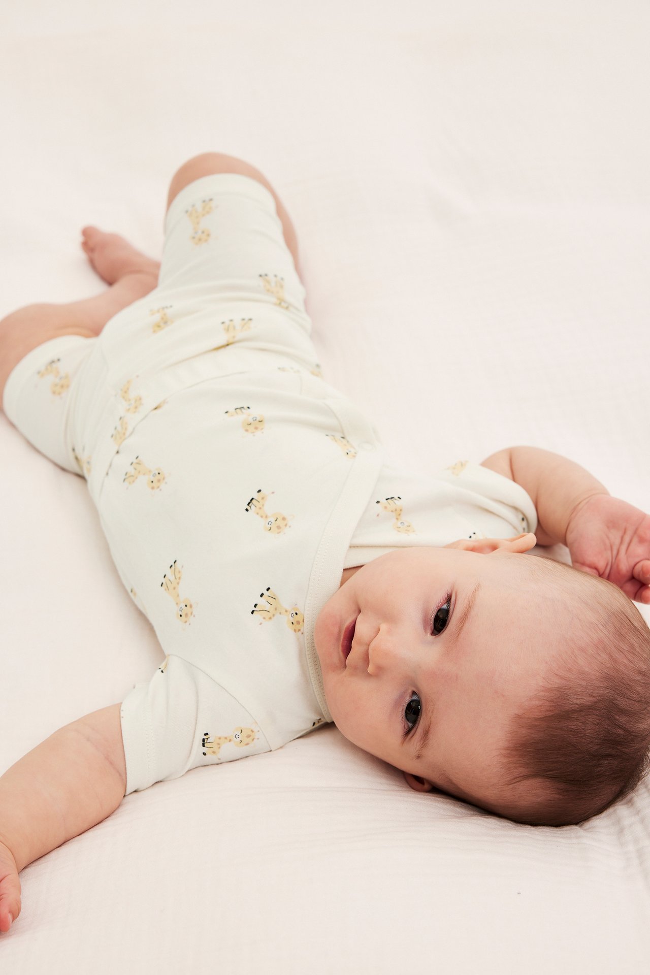 Widok z góry na niemowlę w kremowym, krótkim bawełnianym body z żółtym wzorem żyraf.