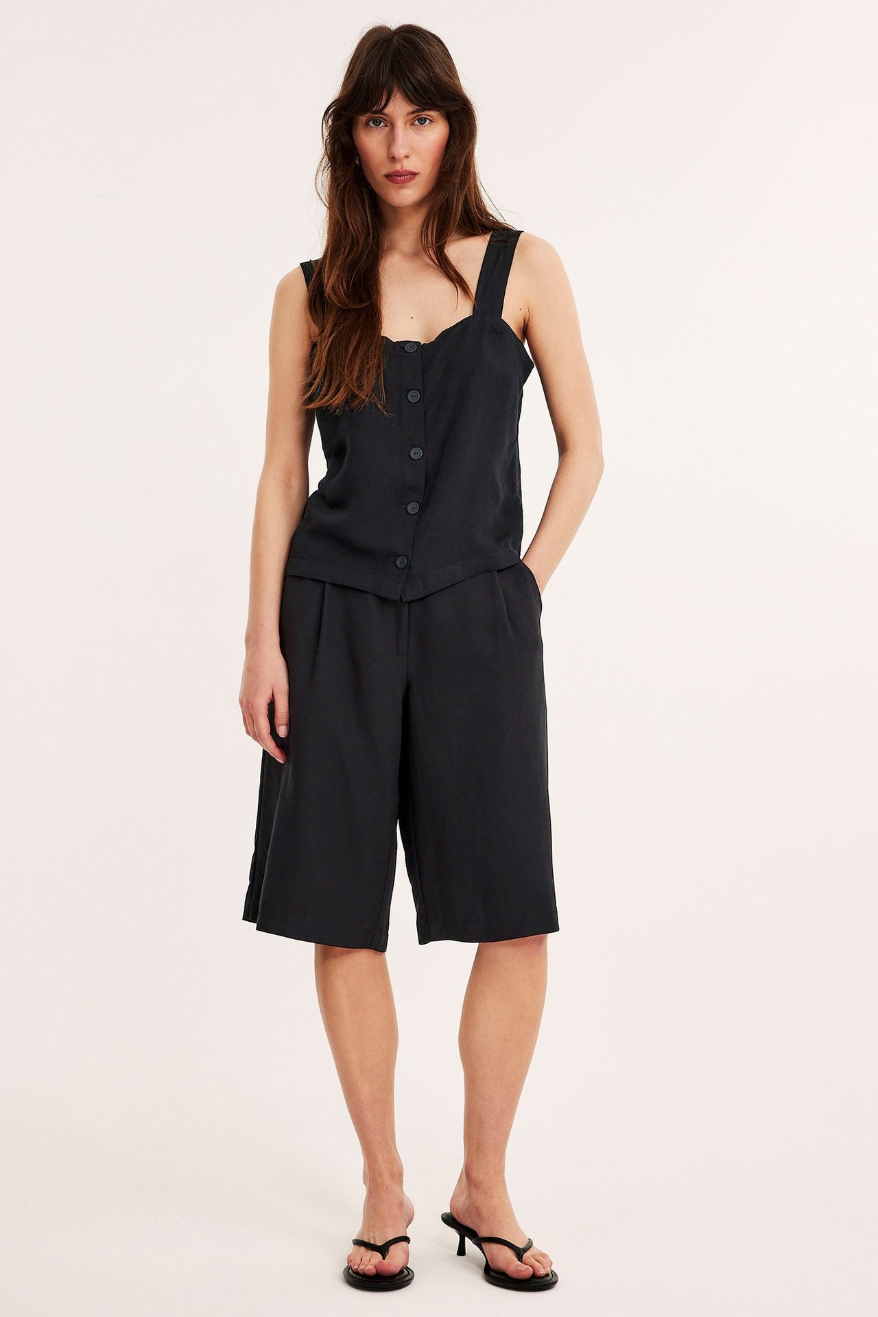 Forfra: kvinne i svart linblandingstopp med knapper, svarte culottes og svarte flip-flops med hæl.