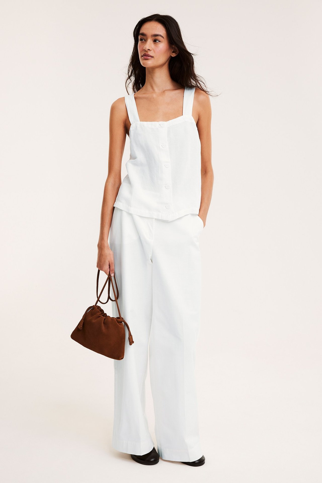 Nainen edestäpäin, yllään valkoinen pellavasekoitetoppi nappikiinnityksellä, valkoiset leveät housut, kädessä ruskea laukku.