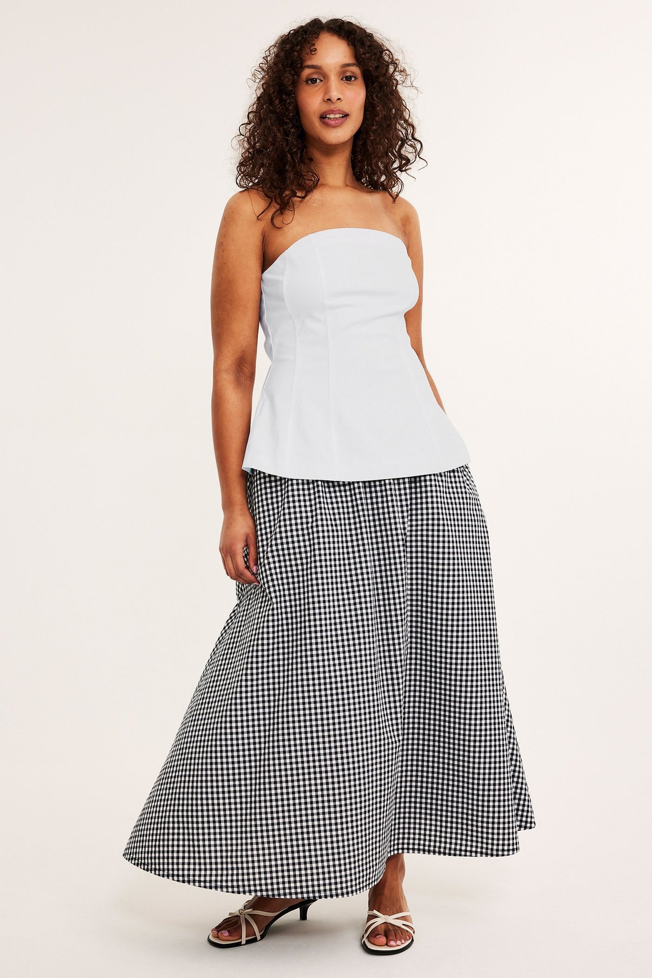 Frontbilde: kvinne i hvit stroppeløs bustier, svart-hvitt rutete maxiskjørt og lyse sandaler.
