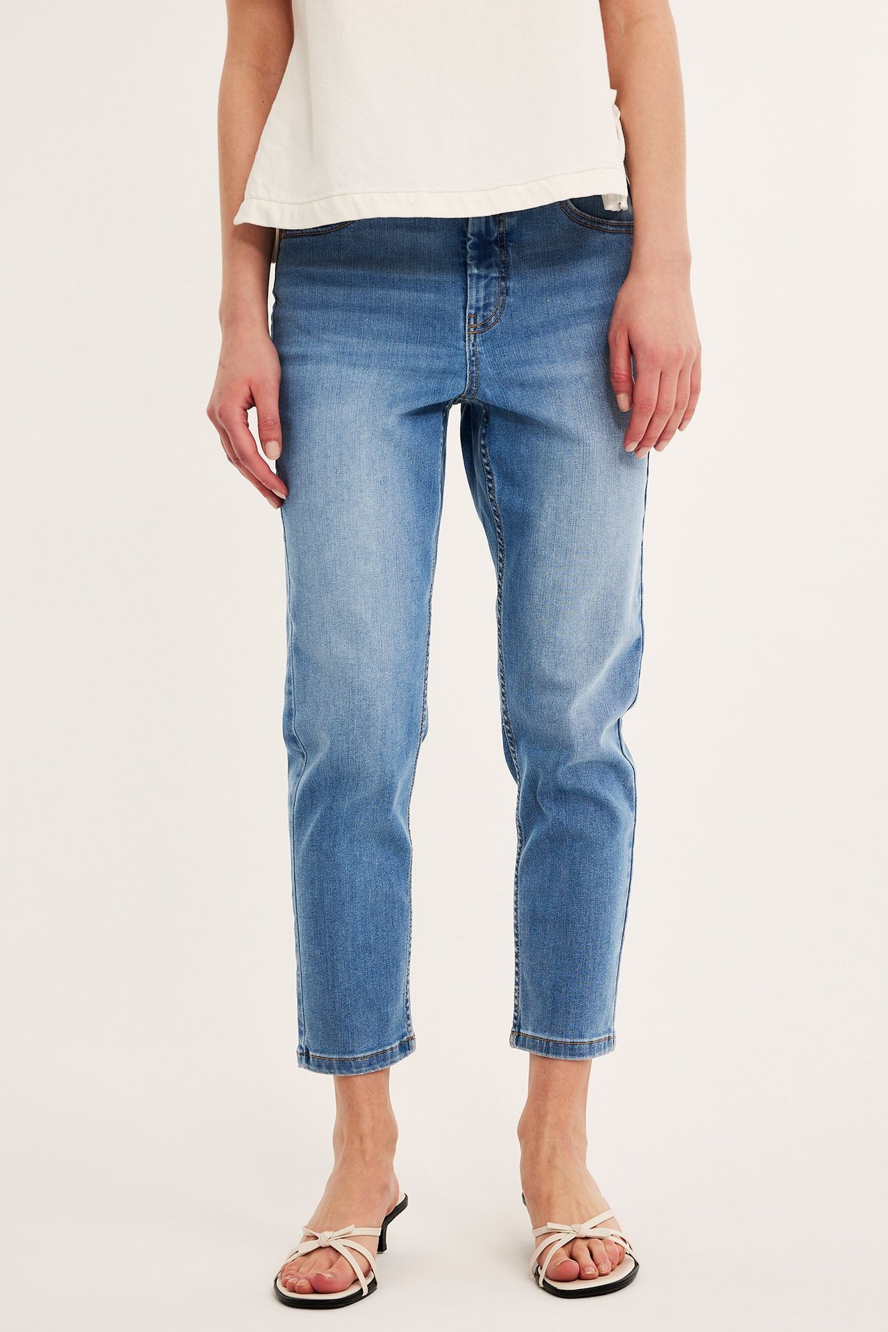 Framifrån, kvinna i ljusblå croppade stretchjeans med hög midja, vit t-shirt och vita klackade sandaler.