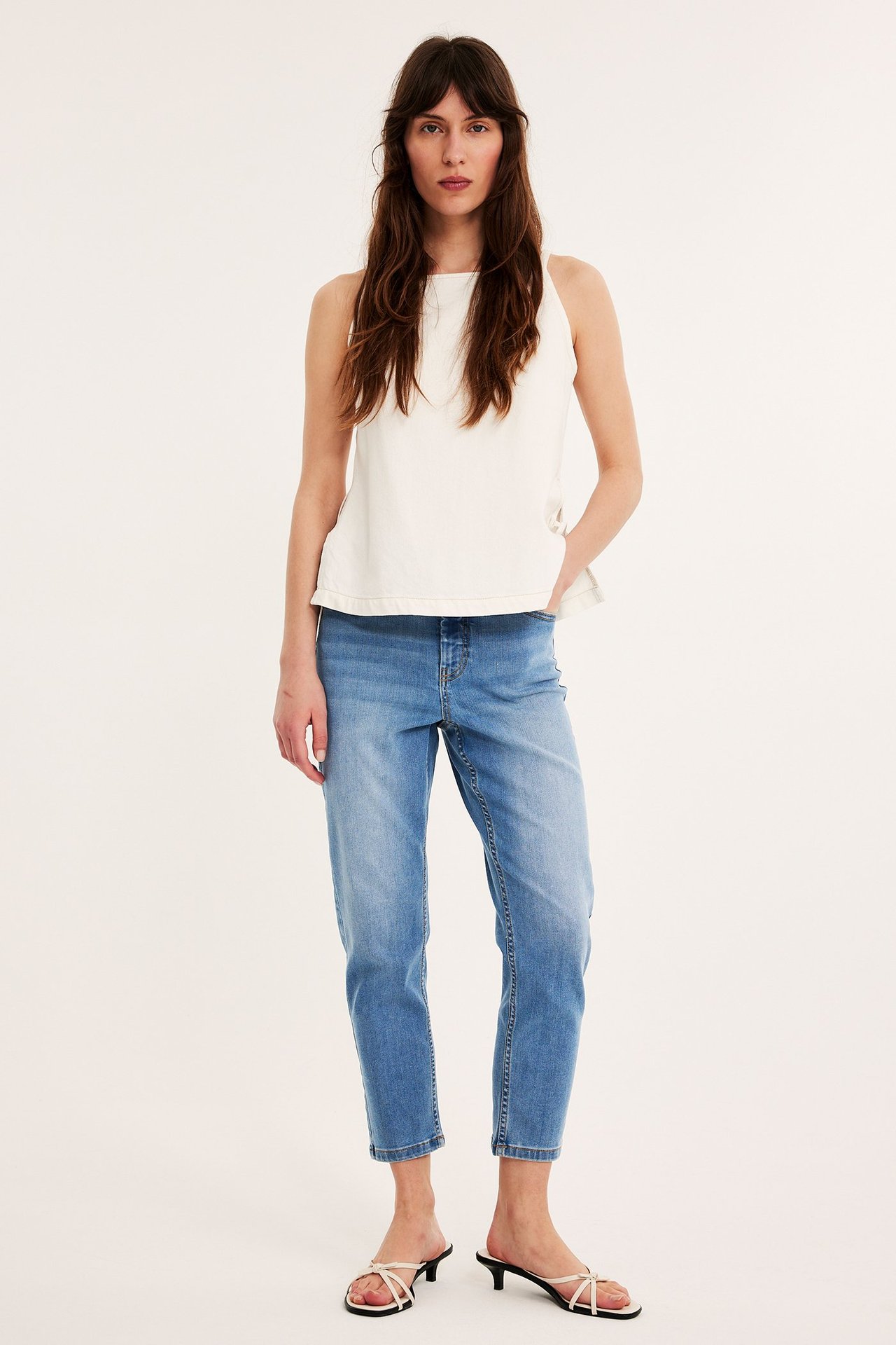Framifrån: Kvinna i ljusblå höga caprijeans i denim, vit ärmlös topp och vita lågklackade sandaler.