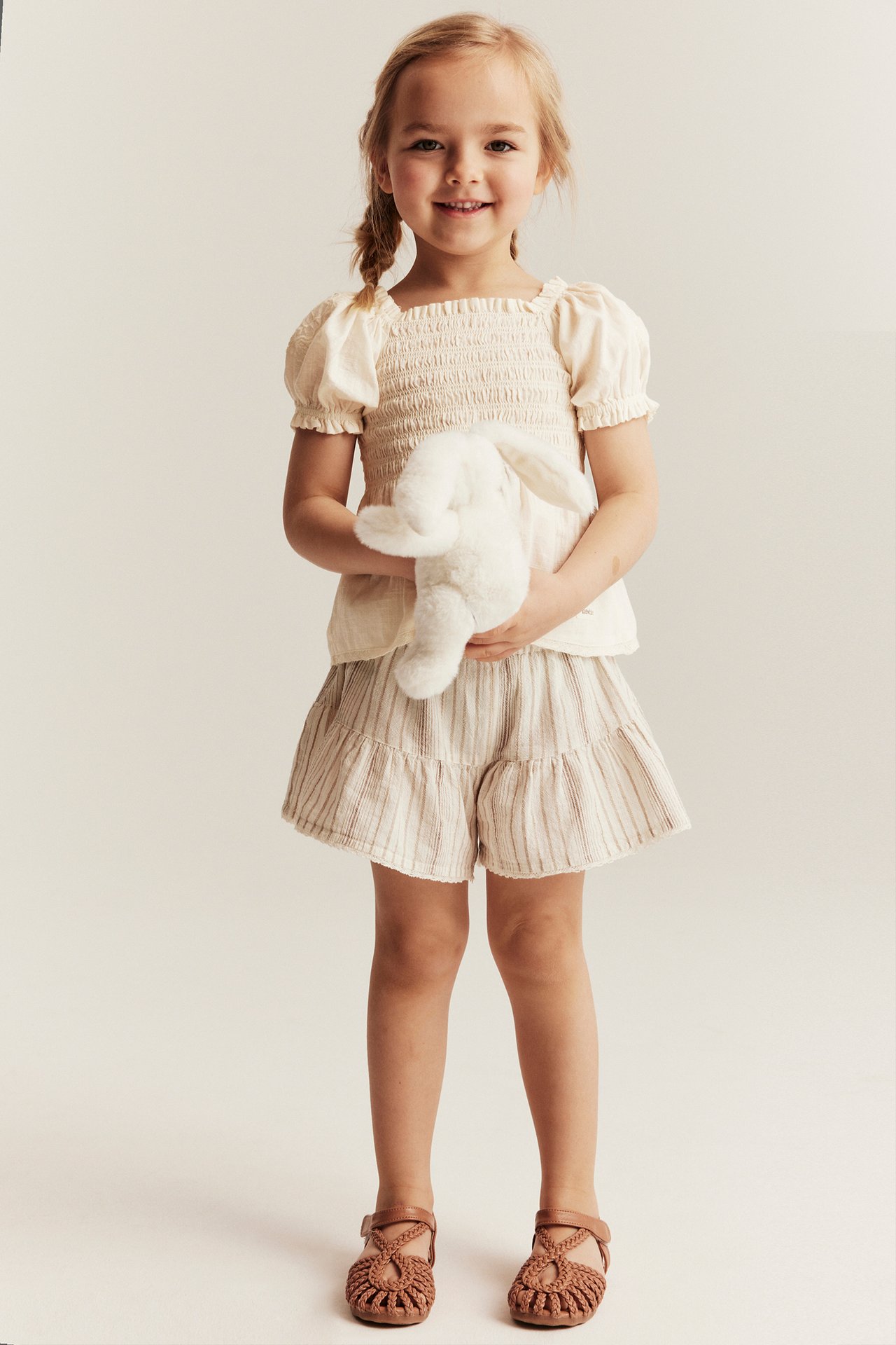 Framifrån: Barn i offwhite smockad bomullsblus, randiga shorts, bruna sandaler, håller vit kanin.