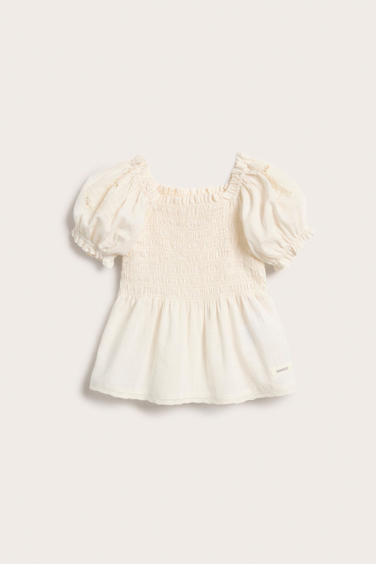 Framifrån, en offwhite bomullsblus för barn med smock, broderade puffärmar och spetskantad peplumfåll.