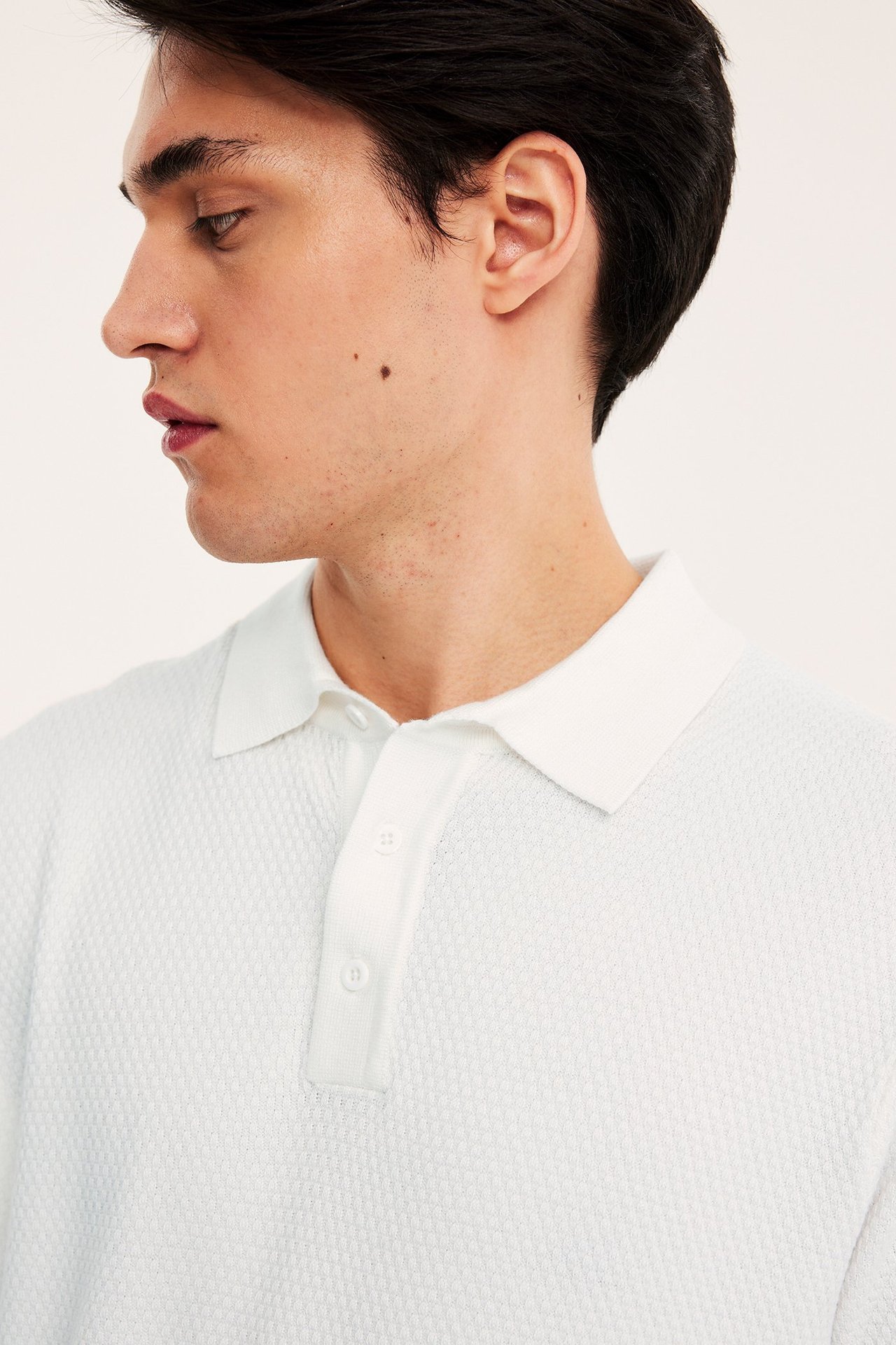 Lähikuva, sivukuva miehestä yllään luonnonvalkoinen neulottu pikeepaita, jossa on rakenne ja kolme nappia.