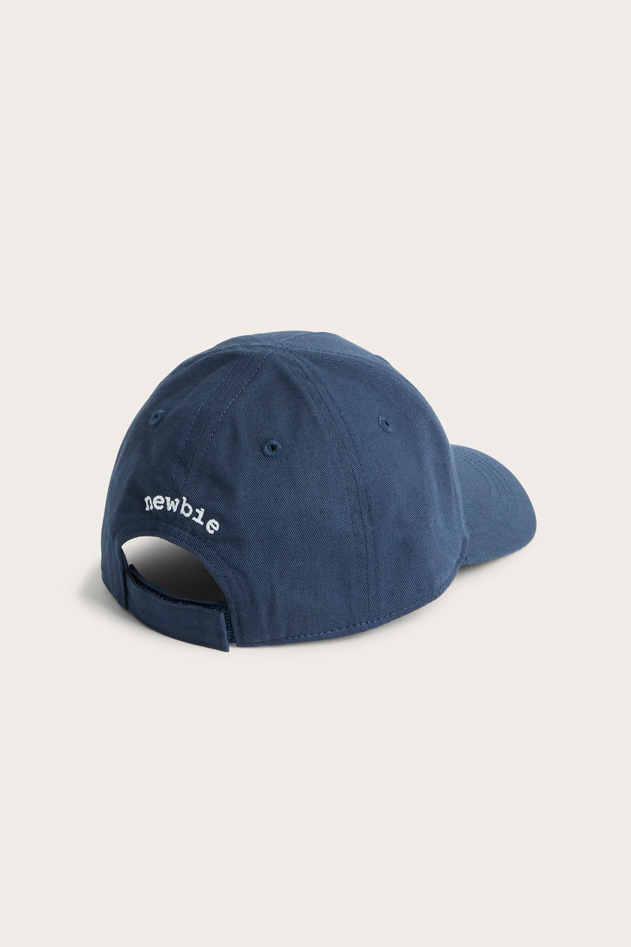 Bakside av en mørkeblå bomullskaps for barn med hvit "newbie" broderi og justerbar stropp.
