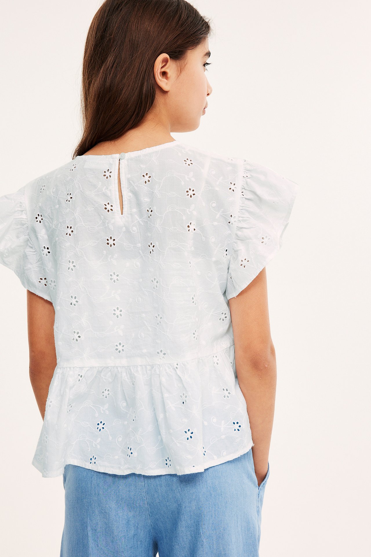 Bakifrån: ett barn i vit bomulls peplumblus med broderie anglaise och ljusblå byxor.