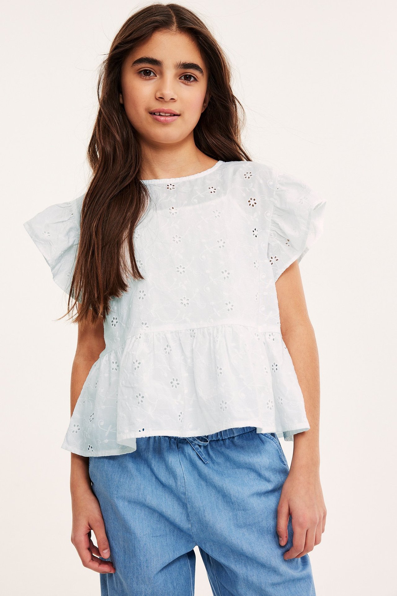 Framifrån: ett barn i ljusblå bomullsblus med peplum och broderie anglaise, samt blåa byxor.