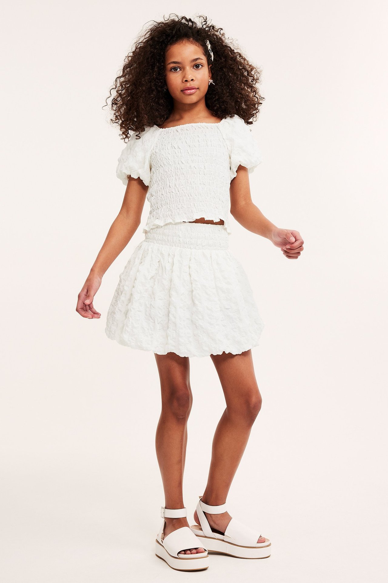 Frontvy: barn i vit strukturerad ballongkjol med smockmidja, matchande smocktopp och vita platåsandaler.