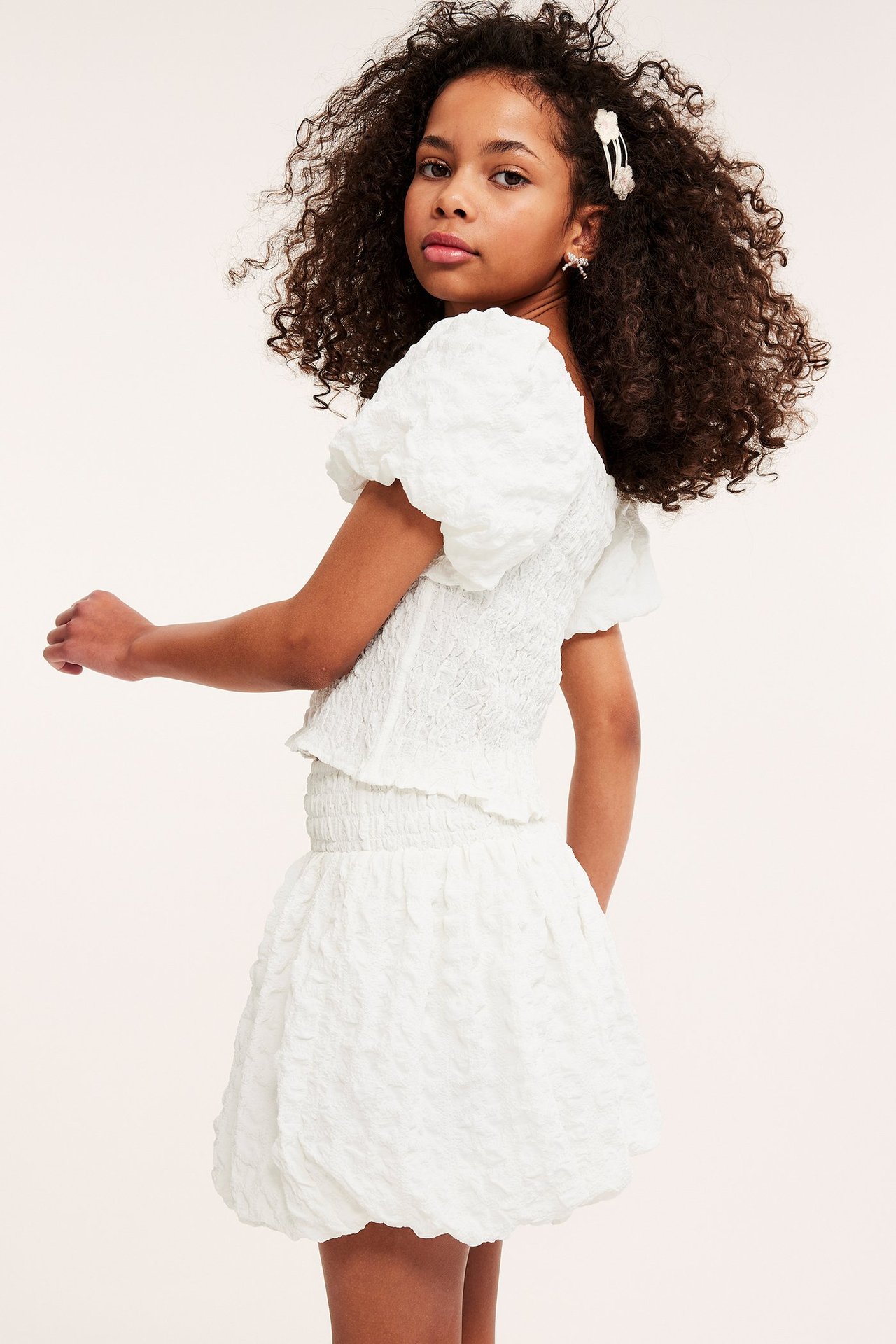 Sidovy av ett barn i vit strukturerad polyester ballongkjol med smock i midjan och matchande topp.