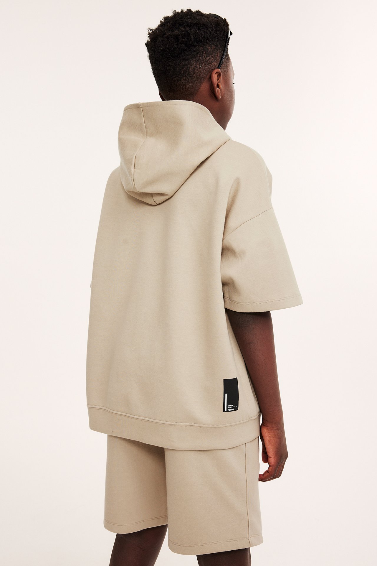 Bakfra av et barn i en beige kortermet interlock-hettegenser og matchende shorts.