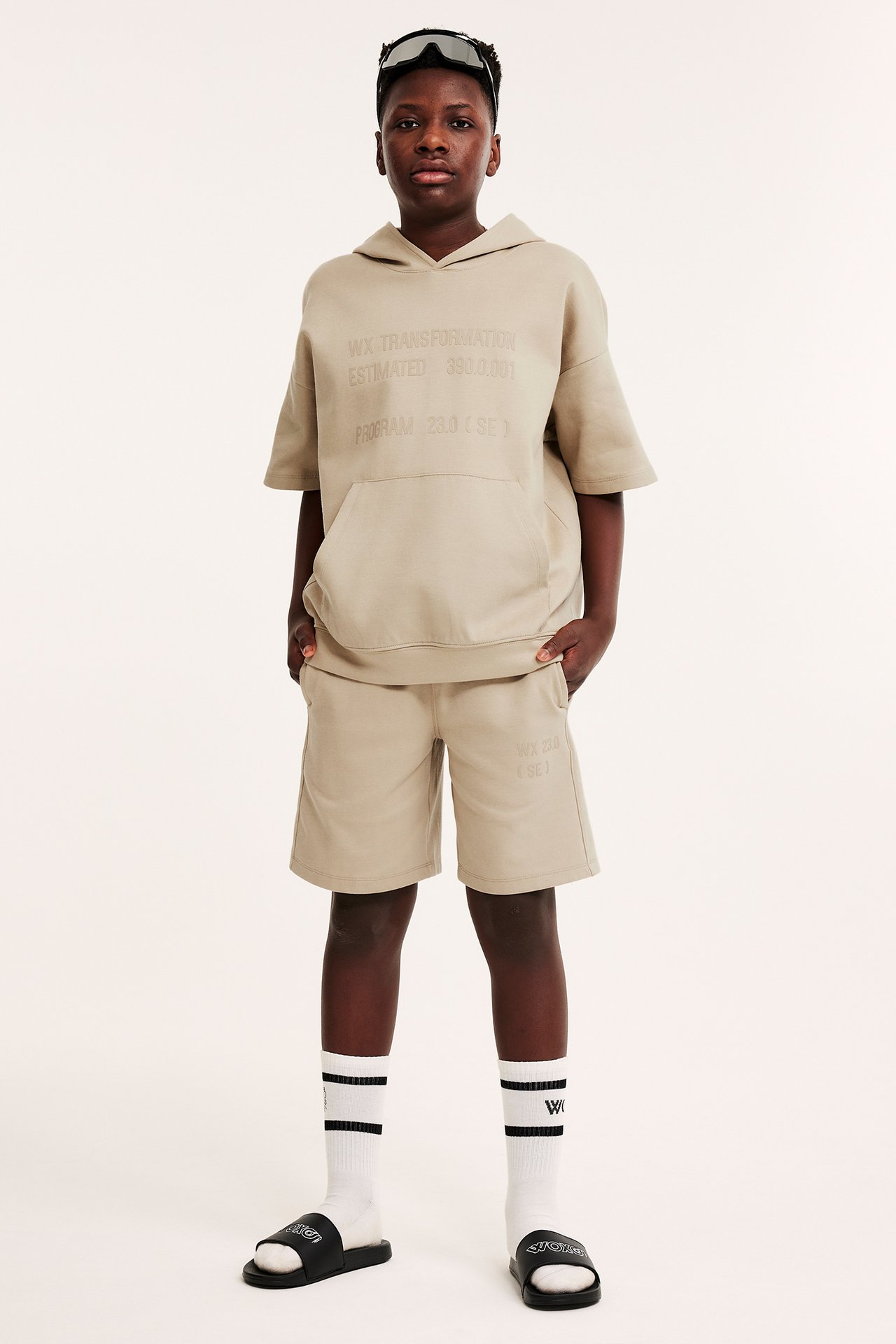 Frontvisning av et barn i beige kortermet hettegenser med trykk, shorts, hvite sokker, svarte sandaler og briller.
