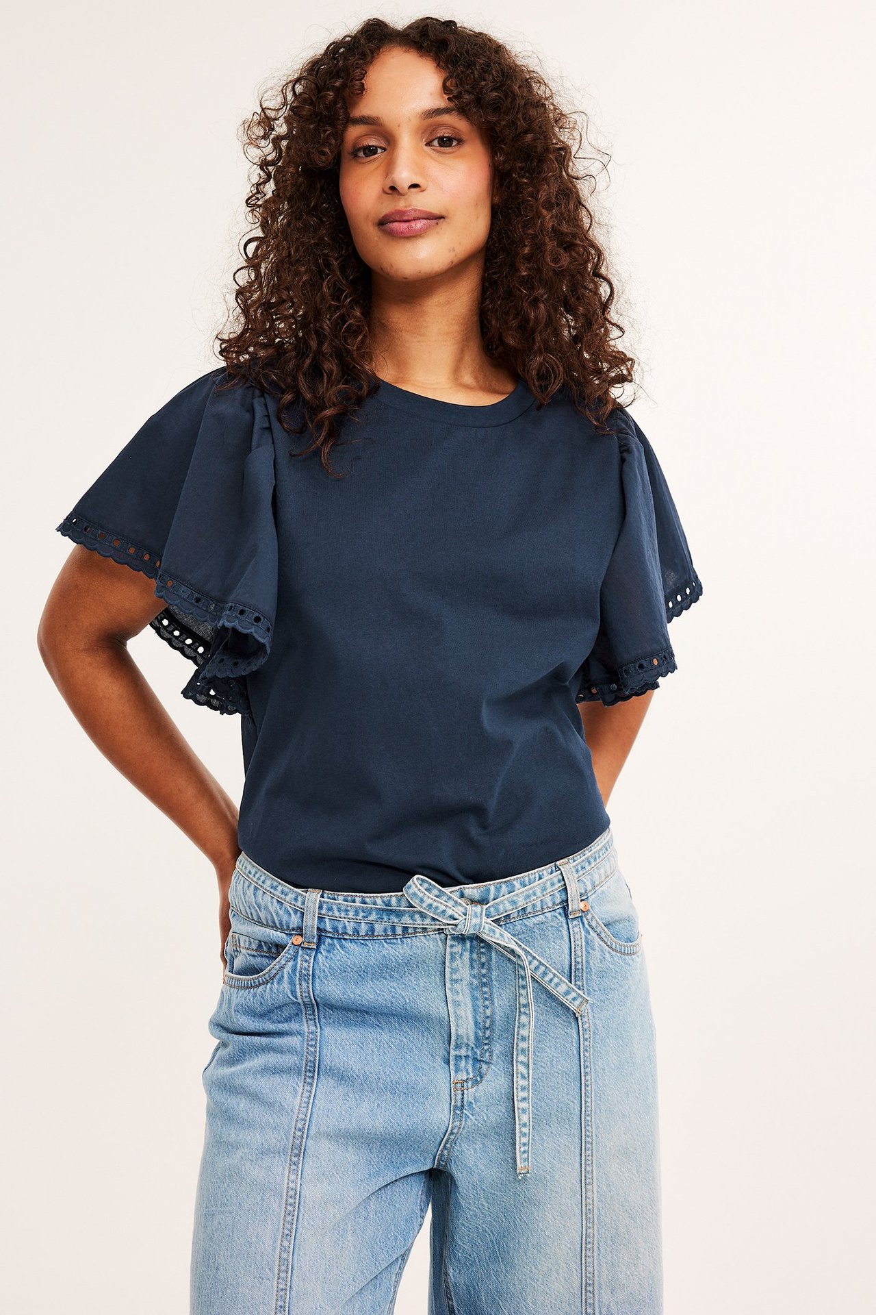 Widok z przodu: kobieta w granatowym bawełnianym topie z falbankami, haftem i jasnoniebieskich dżinsach.