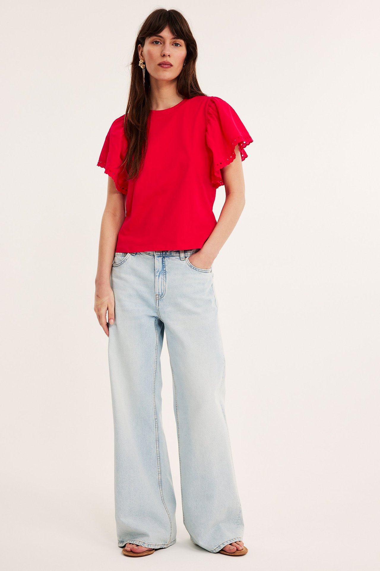Frontvy: Kvinna i röd broderad volangärmad topp och ljusblå vida jeans.