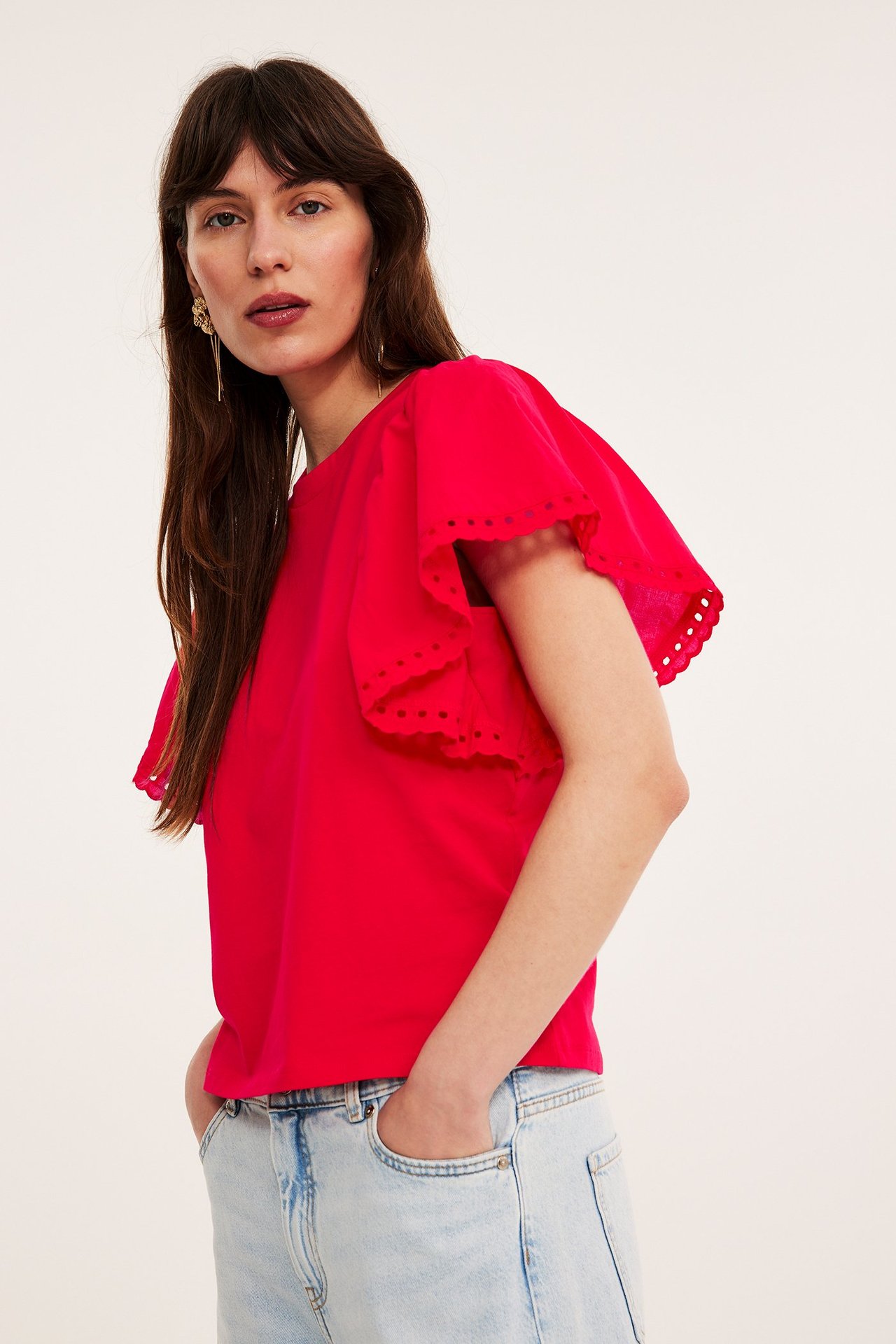 Widok z przodu kobiety w czerwonym bawełnianym topie z haftowanymi falbanami i jasnoniebieskich luźnych jeansach.