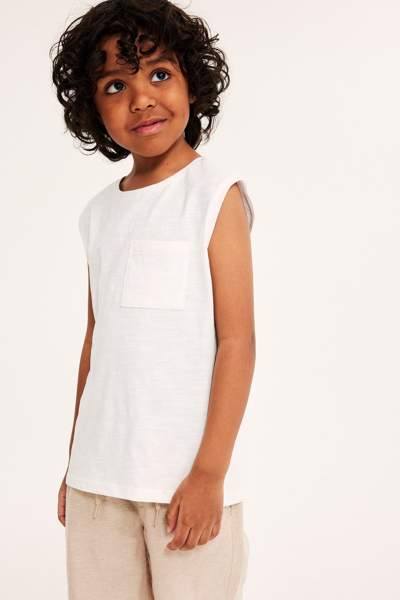 Widok z przodu dziecka w bawełnianym topie bez rękawów w kolorze ecru z kieszonką i beżowych szortach.