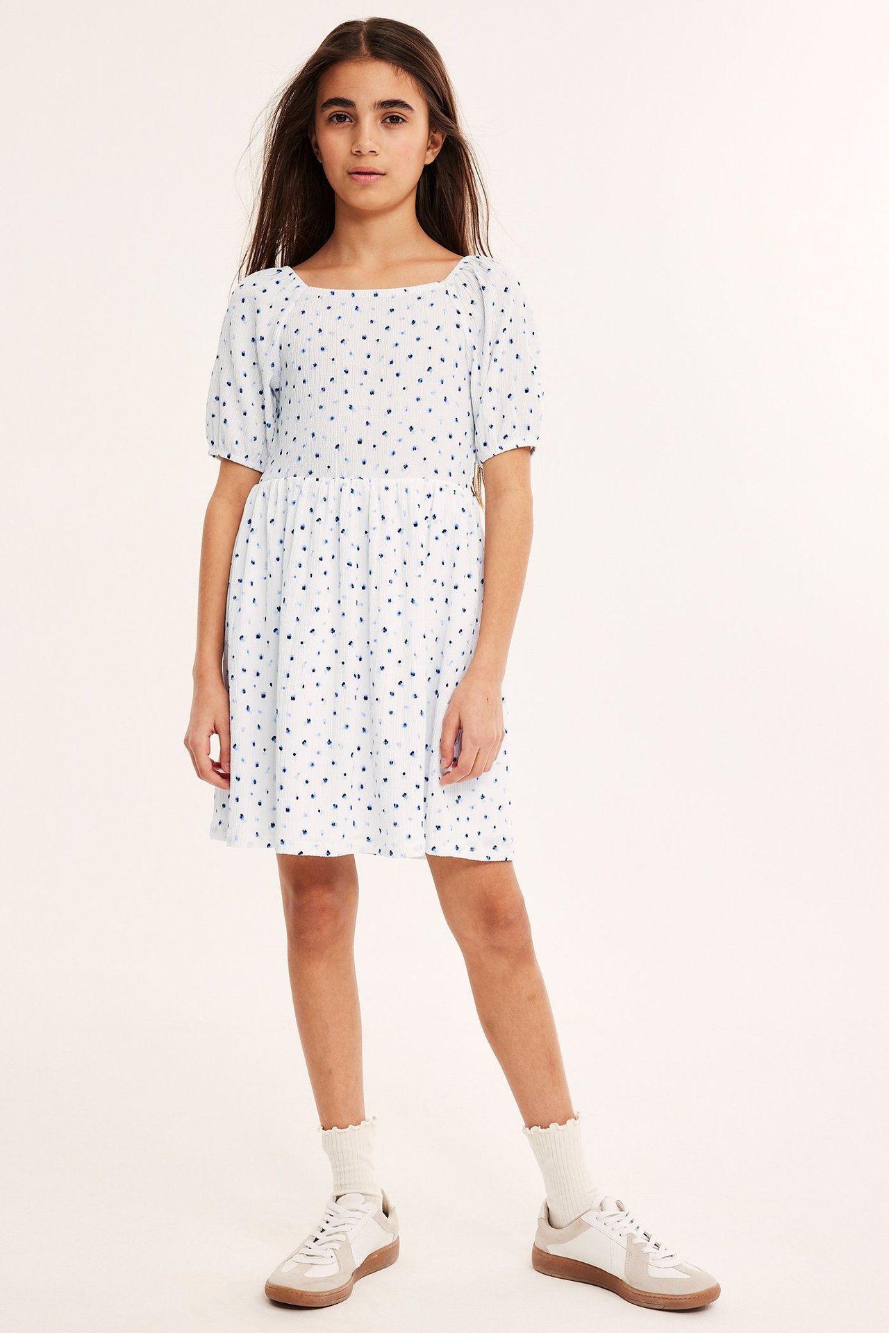 Forfra: Barn i hvit krøllet jerseykjole med blått blomstermønster, hvite rysjesokker og hvite joggesko.