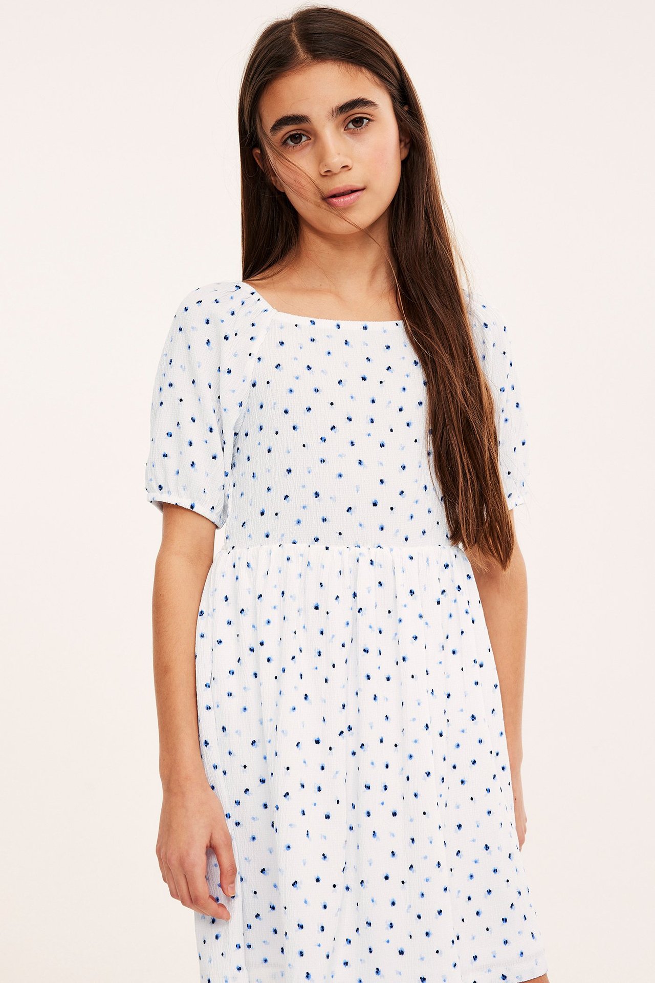 Frontbilde av et barn i hvit krøllet jerseykjole med blått blomstermønster og korte puffermer.