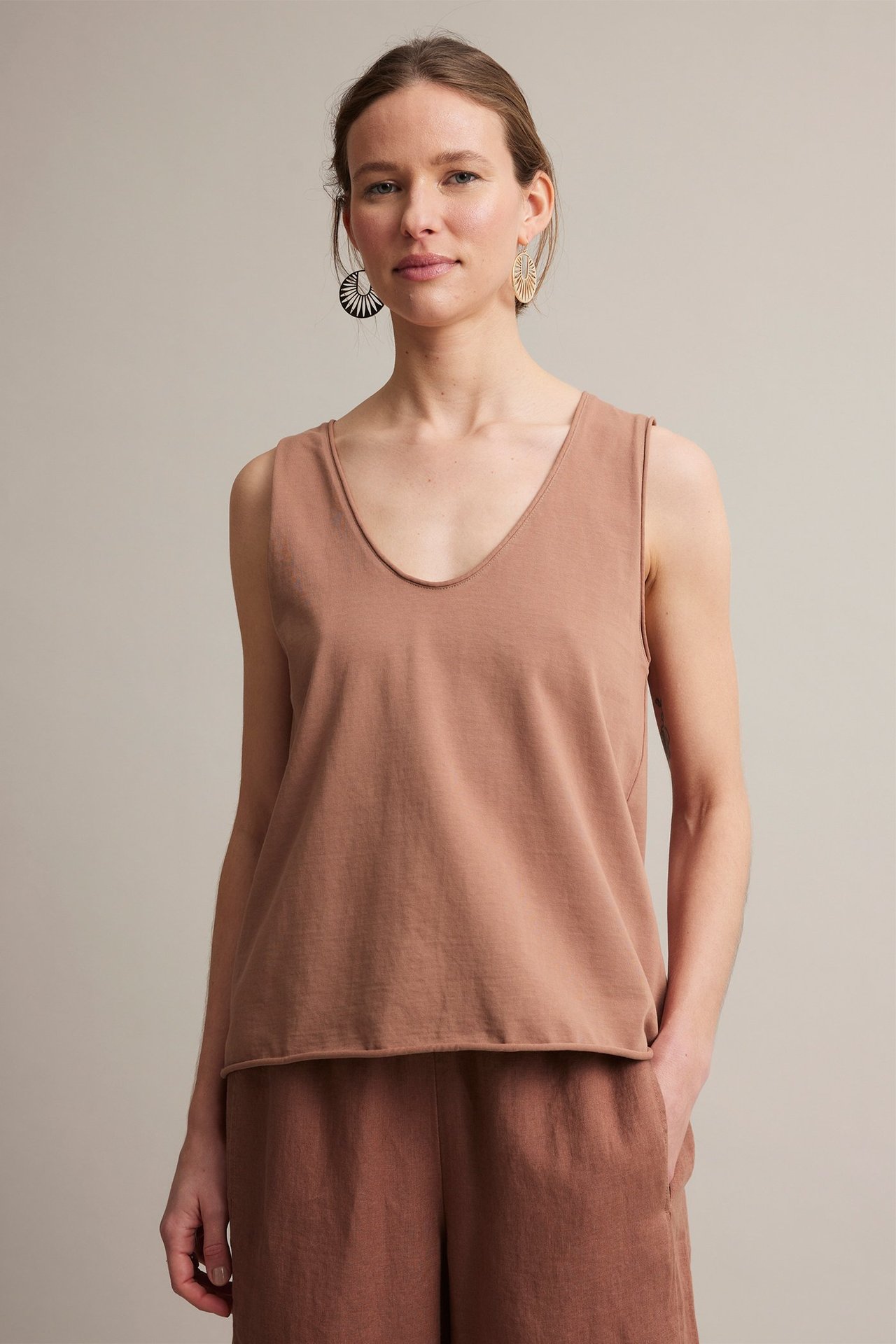 Frontbilde av kvinne i brun bomulls V-hals topp, matchende bukser og mønstrede øredobber.