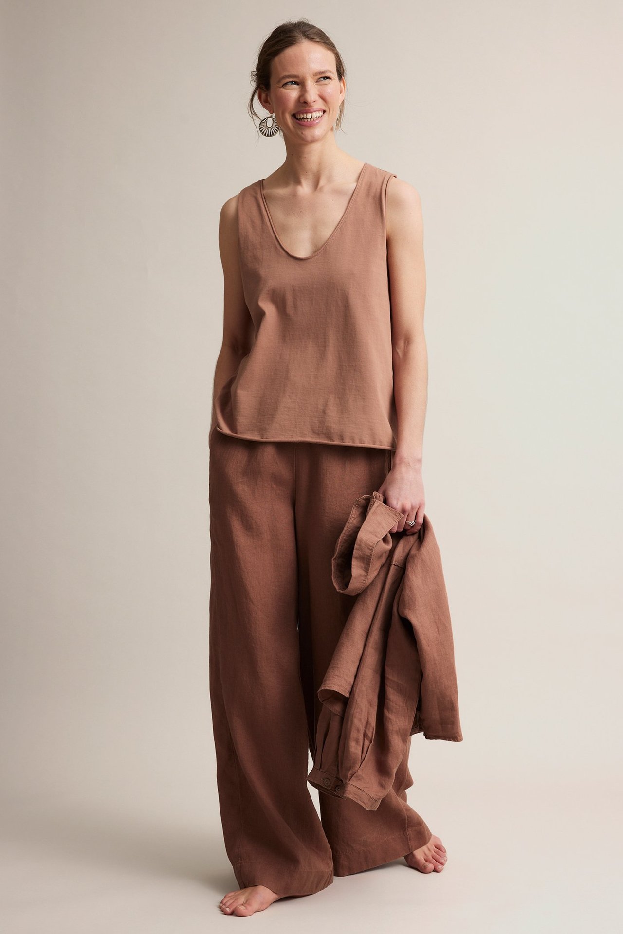 Forfra, kvinne i brun V-hals bomullstopp, matchende vide bukser, holder en brun skjorte.