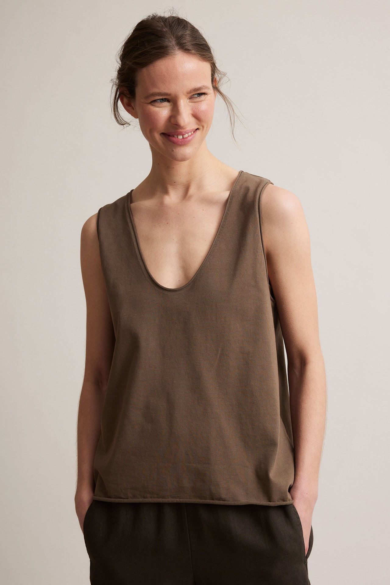 Forfra: kvinne i brun V-hals bomullstopp med rå kanter og mørke bukser.