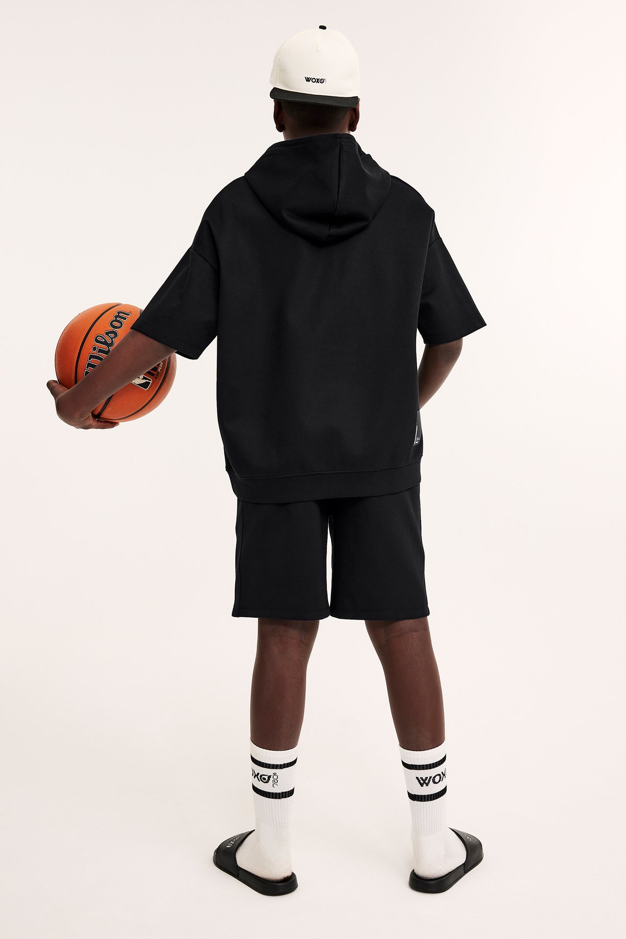 Ryggvy av barn i svarta interlockshorts, huvtröja, vit keps, strumpor och tofflor, hållande en basketboll.