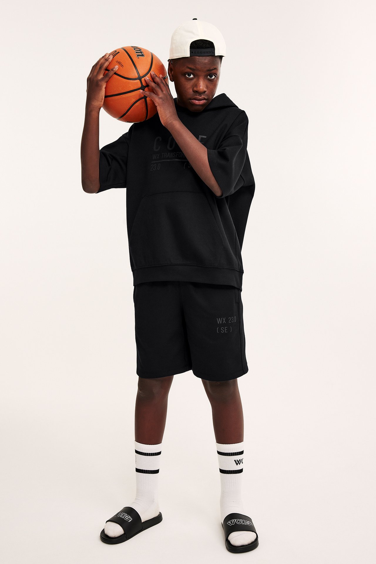 Framifrån: Barn i svarta shorts med tryck, svart huvtröja, vit keps, vita strumpor, svarta tofflor, håller boll.