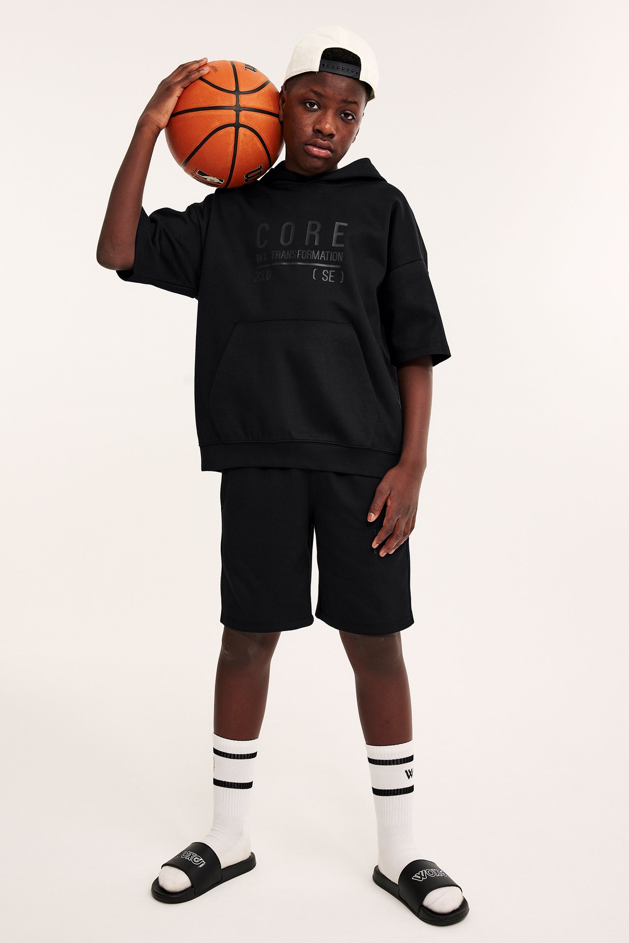 Framifrån: Kid i svarta interlockshorts, svart kortärmad huvtröja, vit keps, strumpor och tofflor, håller en basketboll.