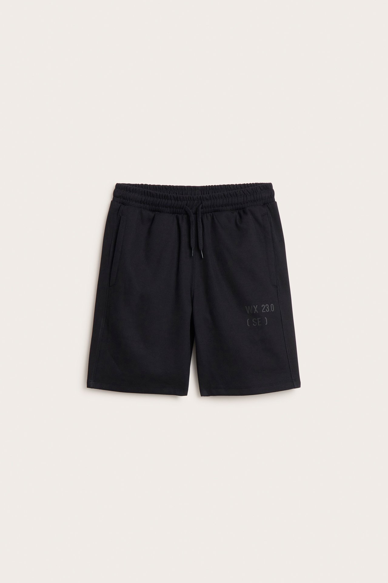 Framifrån, svarta interlock-shorts för barn med resår i midjan, dragsko, sidfickor och mörkt tryck.