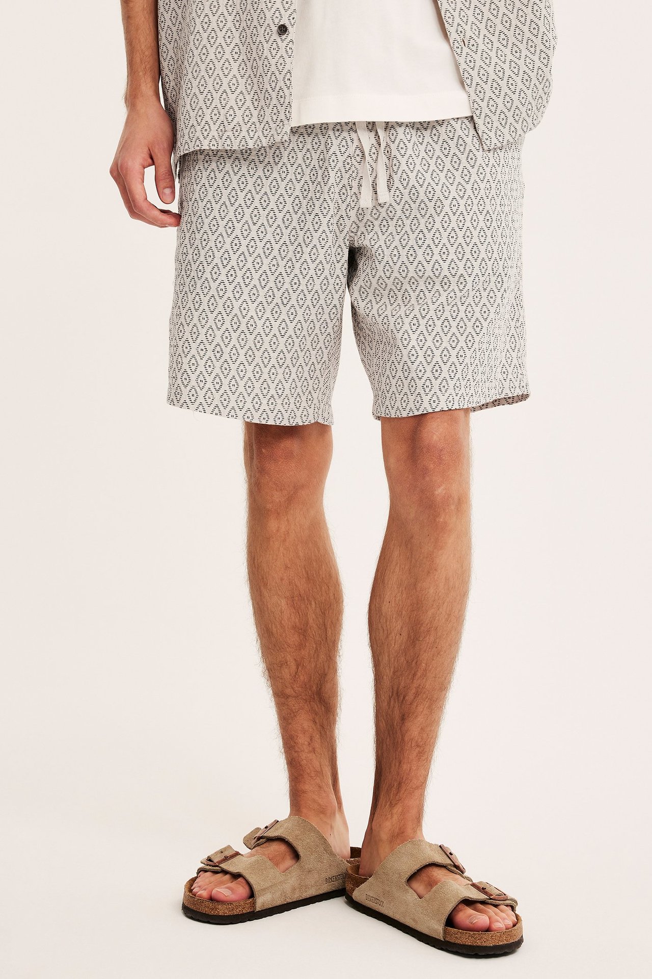 Edestä, mies yllään vaaleanbeiget kuviolliset puuvillashortsit, yhteensopiva paita ja beiget sandaalit.