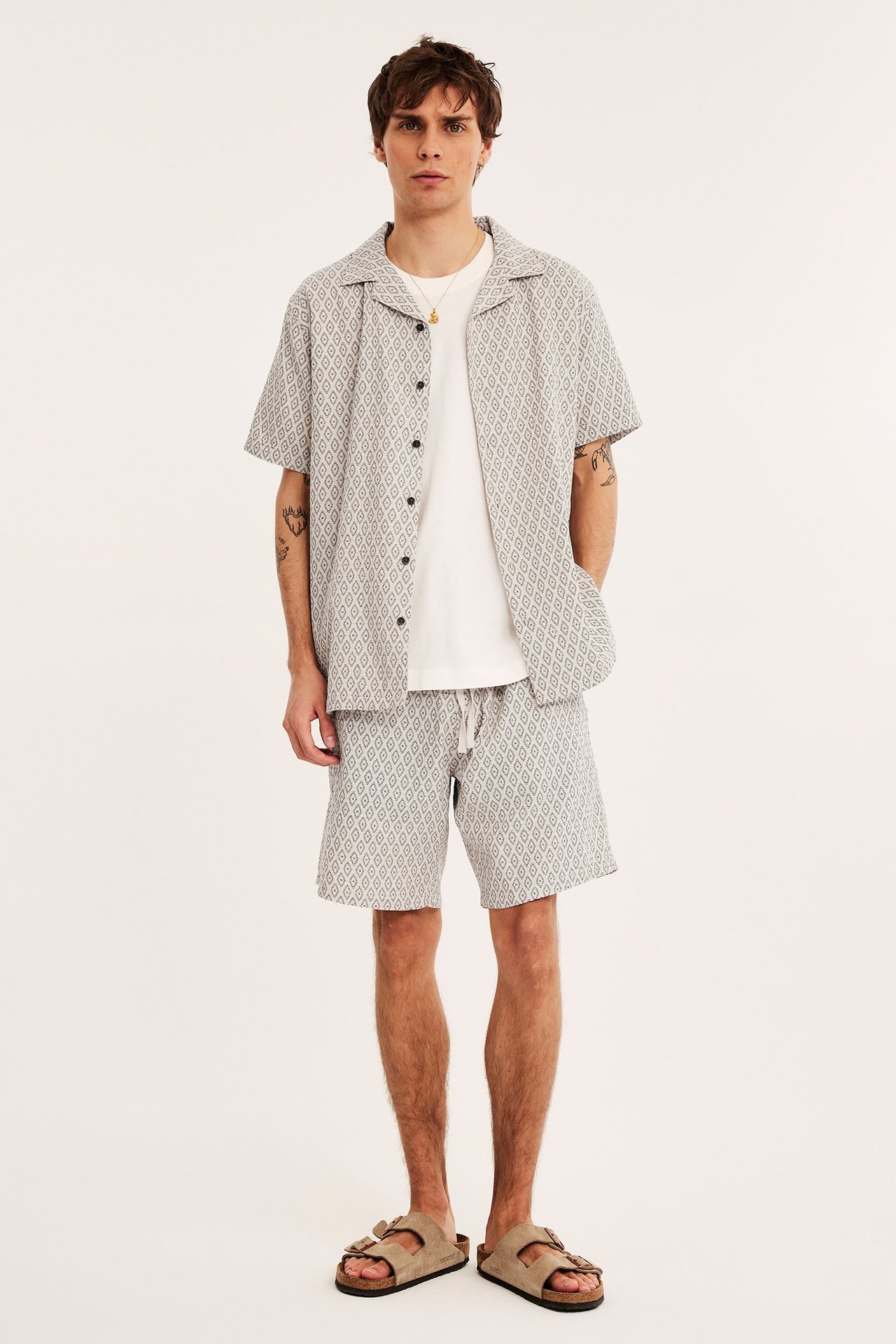 Edestä: mies vaaleanbeiget kuviolliset puuvillashortsit, paita, valkoinen t-paita, beiget sandaalit.