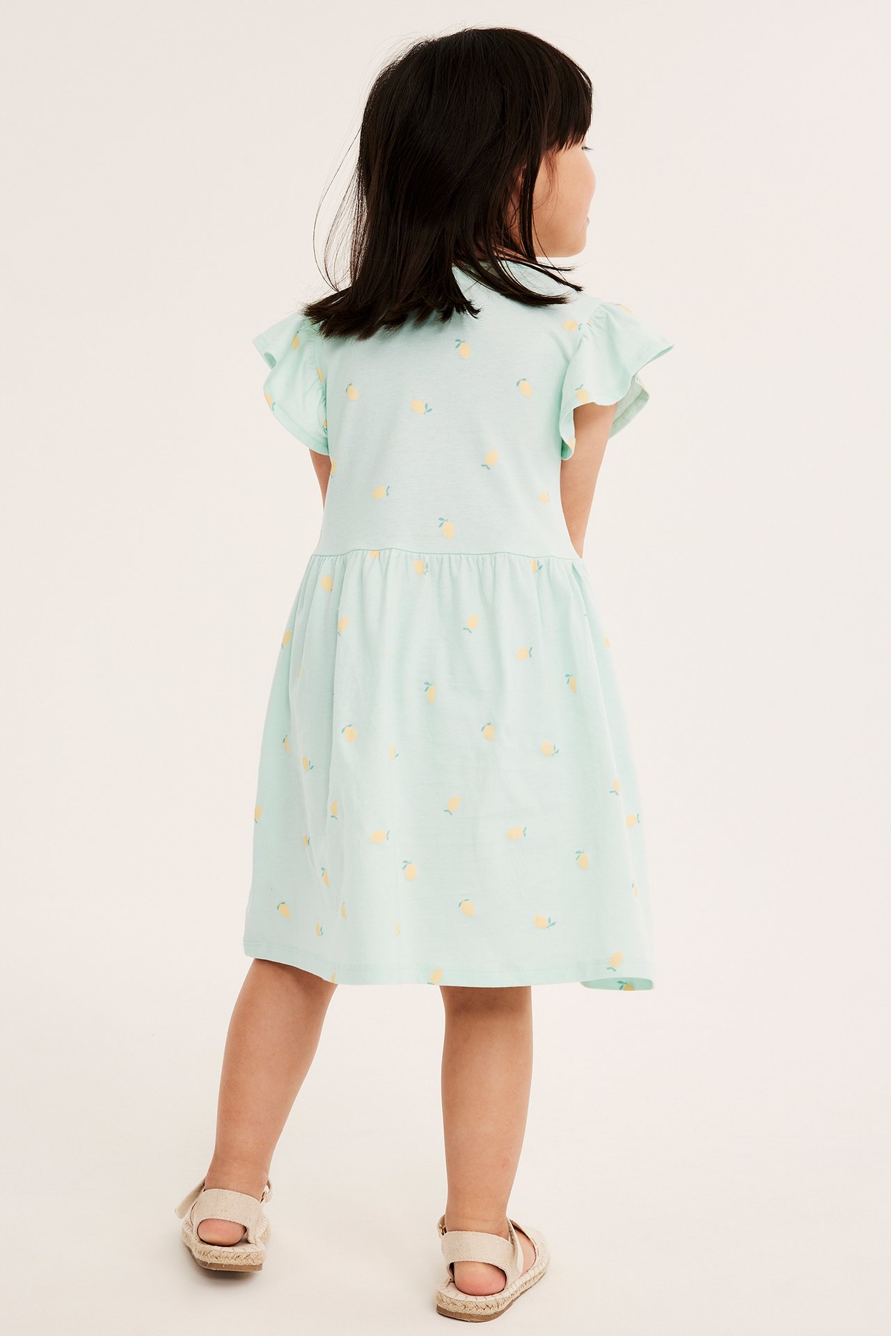 Bakfra, et barn i lysegrønn bomullskjole med sitronmønster, volangermer og beige espadrillesandaler.
