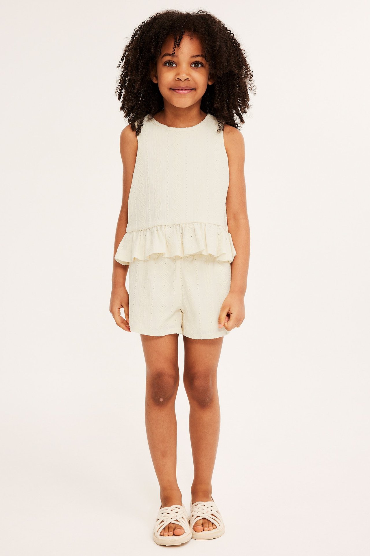 Widok z przodu dziecka w kremowych szortach dżersejowych we wzory, pasującym topie bez rękawów i sandałach.