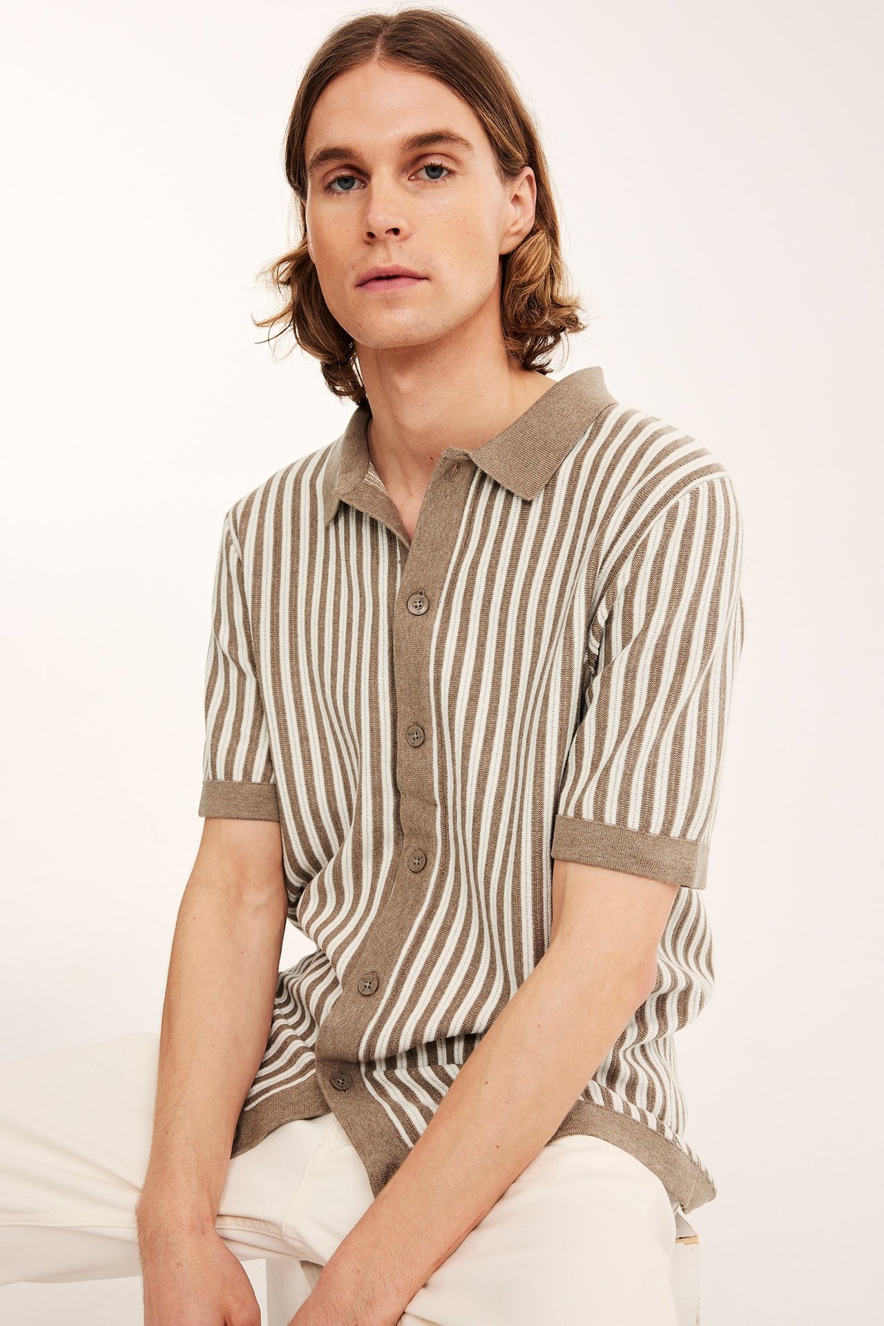 Edestä: mies beigemeleratussa ja valkoisessa raidallisessa neulepolopaidassa, napitettu, vaaleissa housuissa.