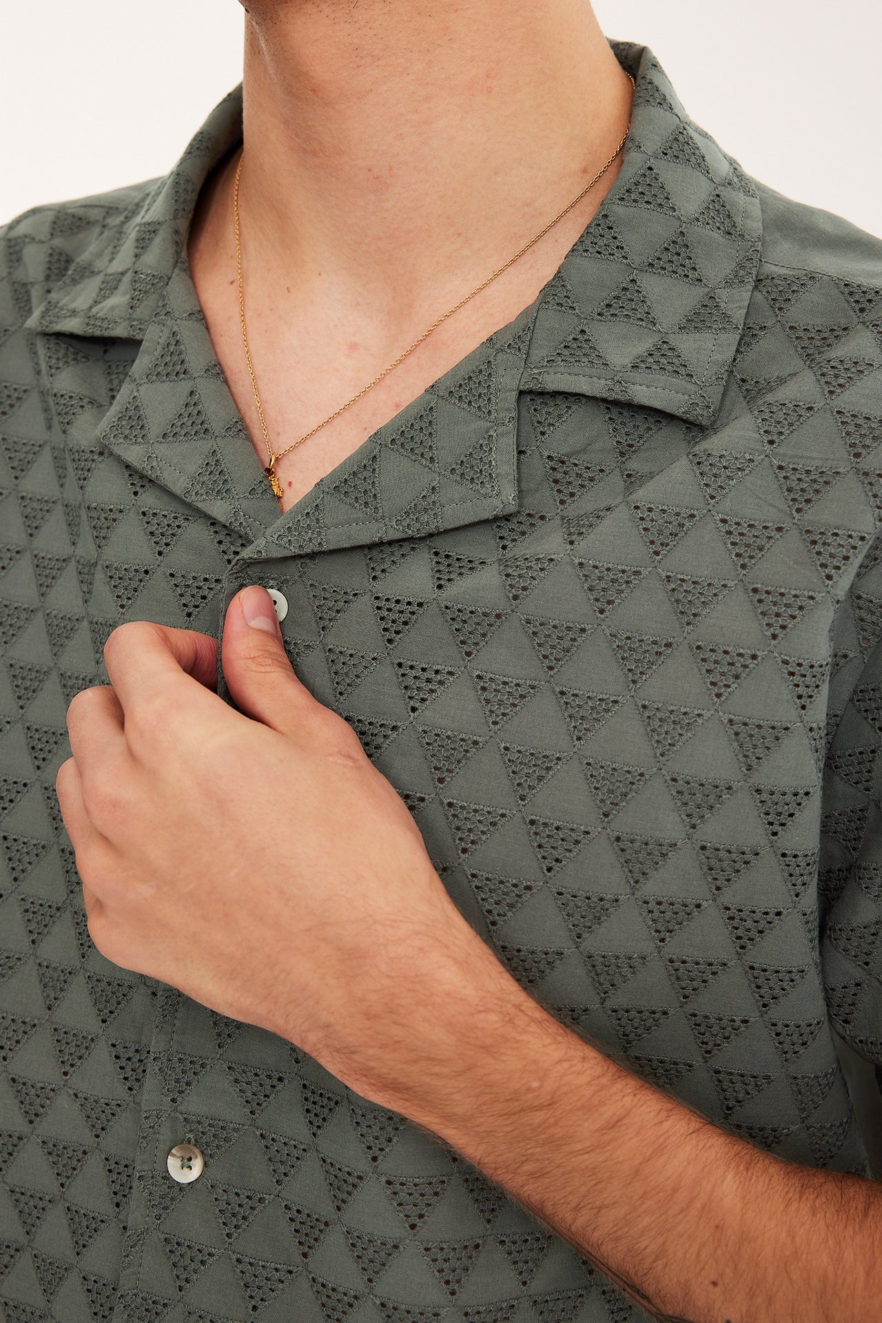 Nærbilde av en mann i grønn kortermet bomullsskjorte med geometrisk broderie anglaise og et gullkjede.