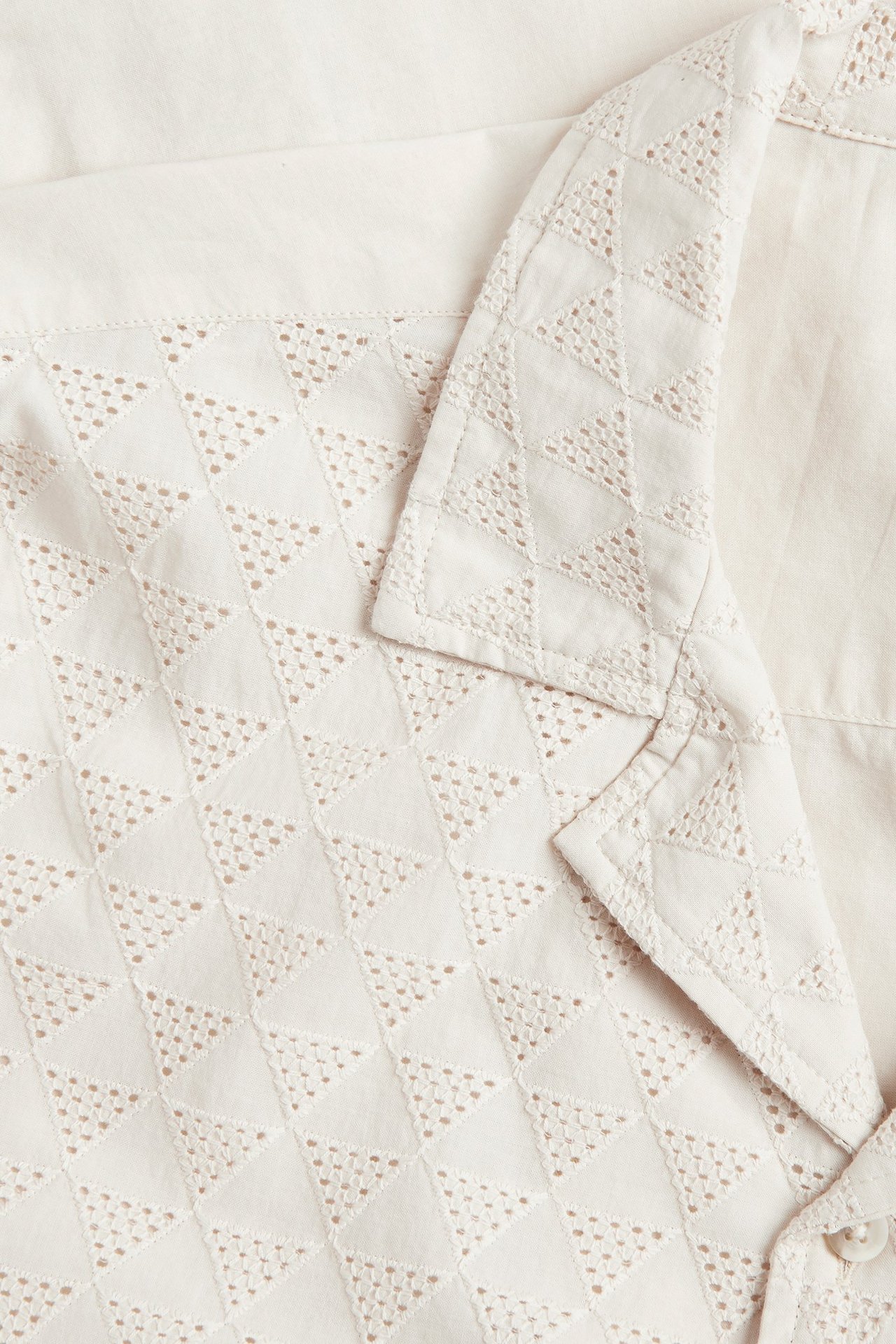 Zbliżenie jasnobeżowej bawełnianej koszuli z krótkim rękawem, z trójkątnym wzorem broderie anglaise z przodu.