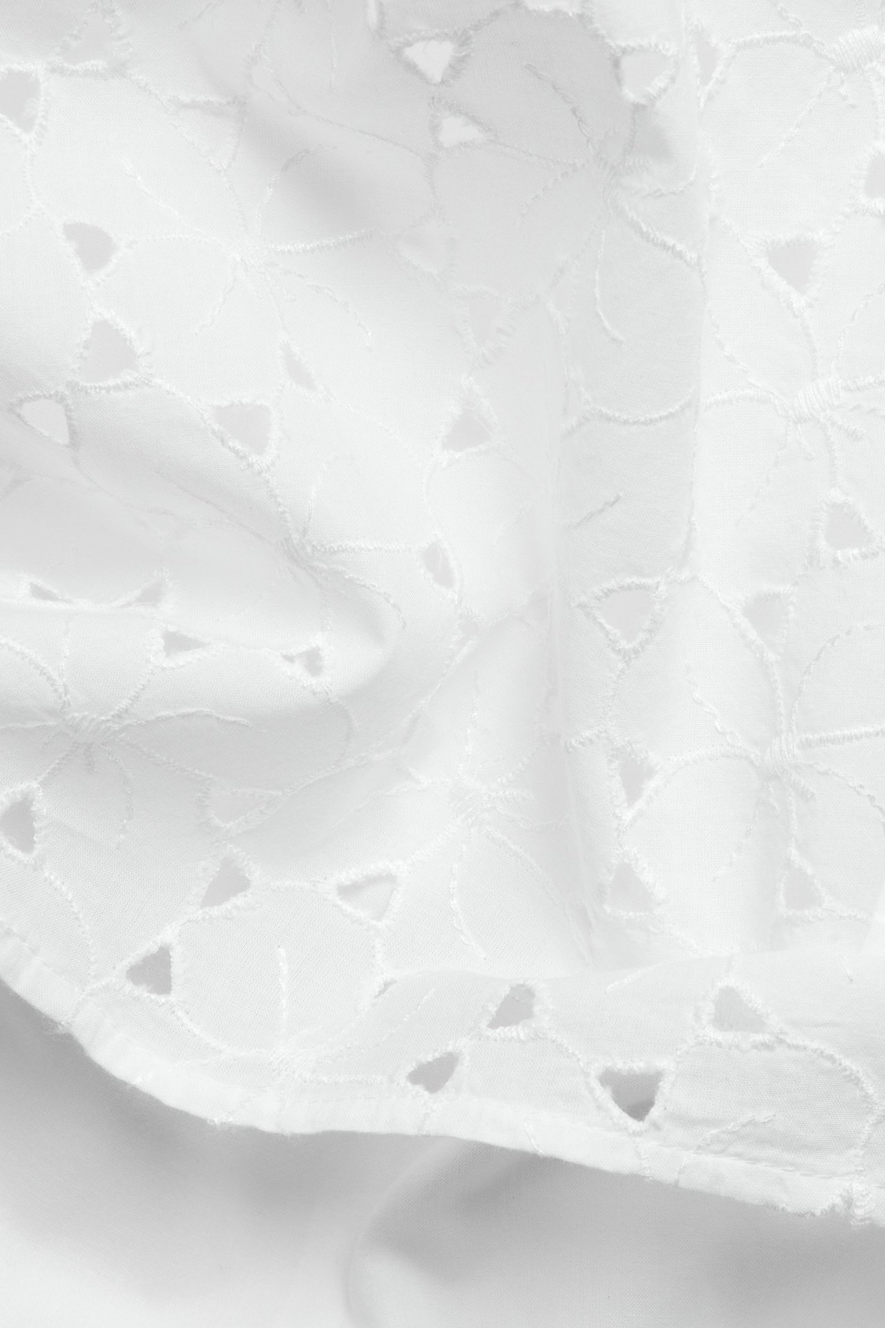 Zbliżenie białej bawełnianej tkaniny z misternymi wycinanymi haftami, tworzącymi delikatny wzór koronki.