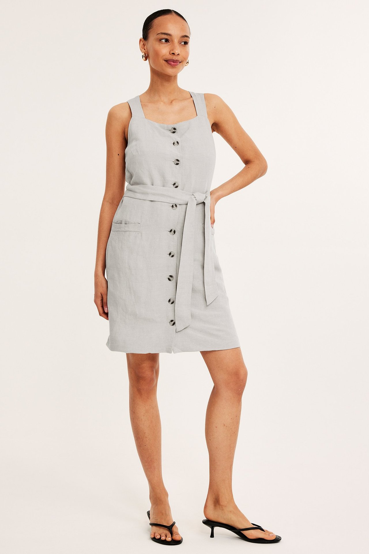 Forfra: Kvinne har på seg lys beige melert linblandingskjole med knapper, knytebelte, svarte sandaler, gulløredobber.