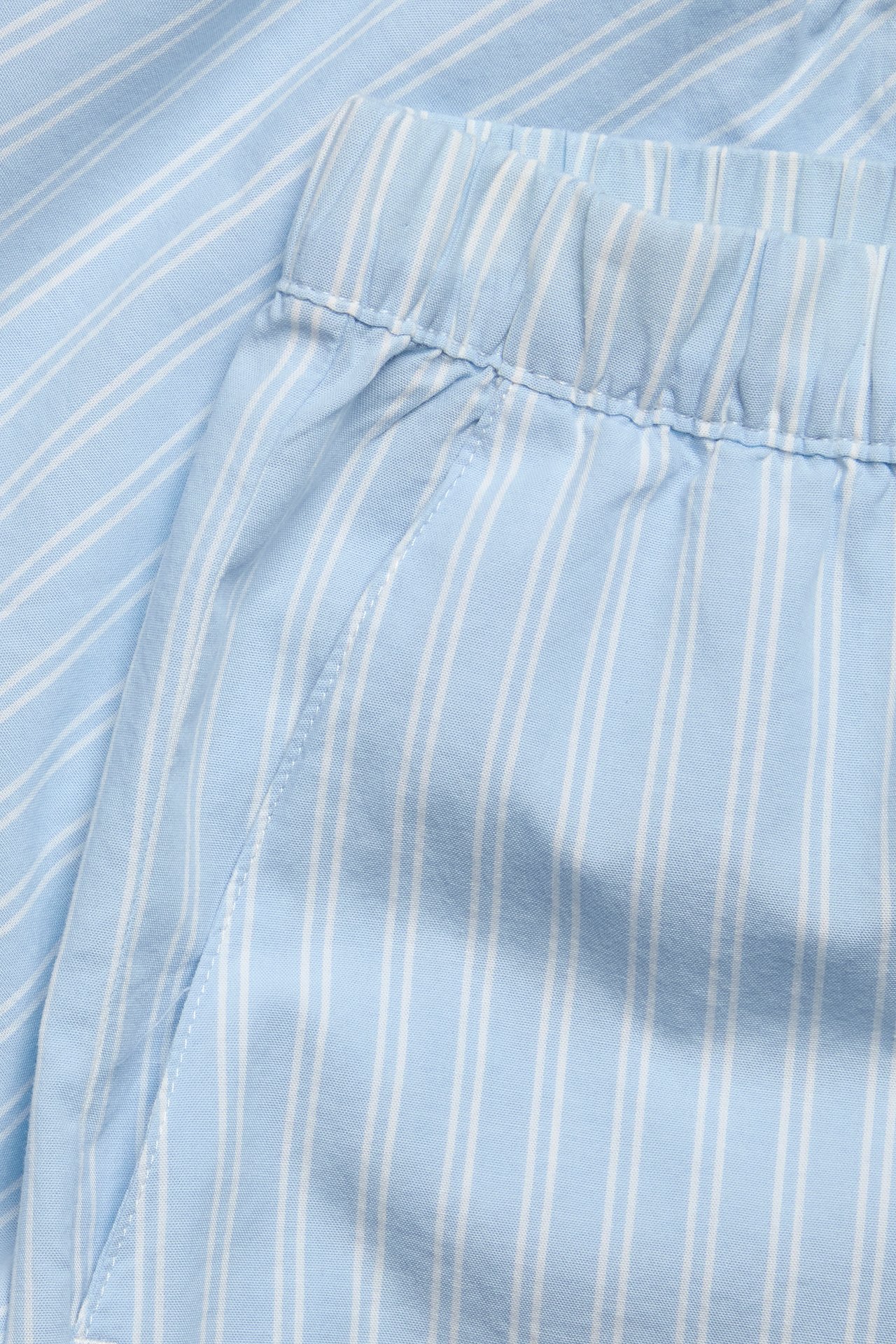 Närbild av ljusblå och vitrandiga pyjamasshorts i bomull med resår i midjan och sidficka.