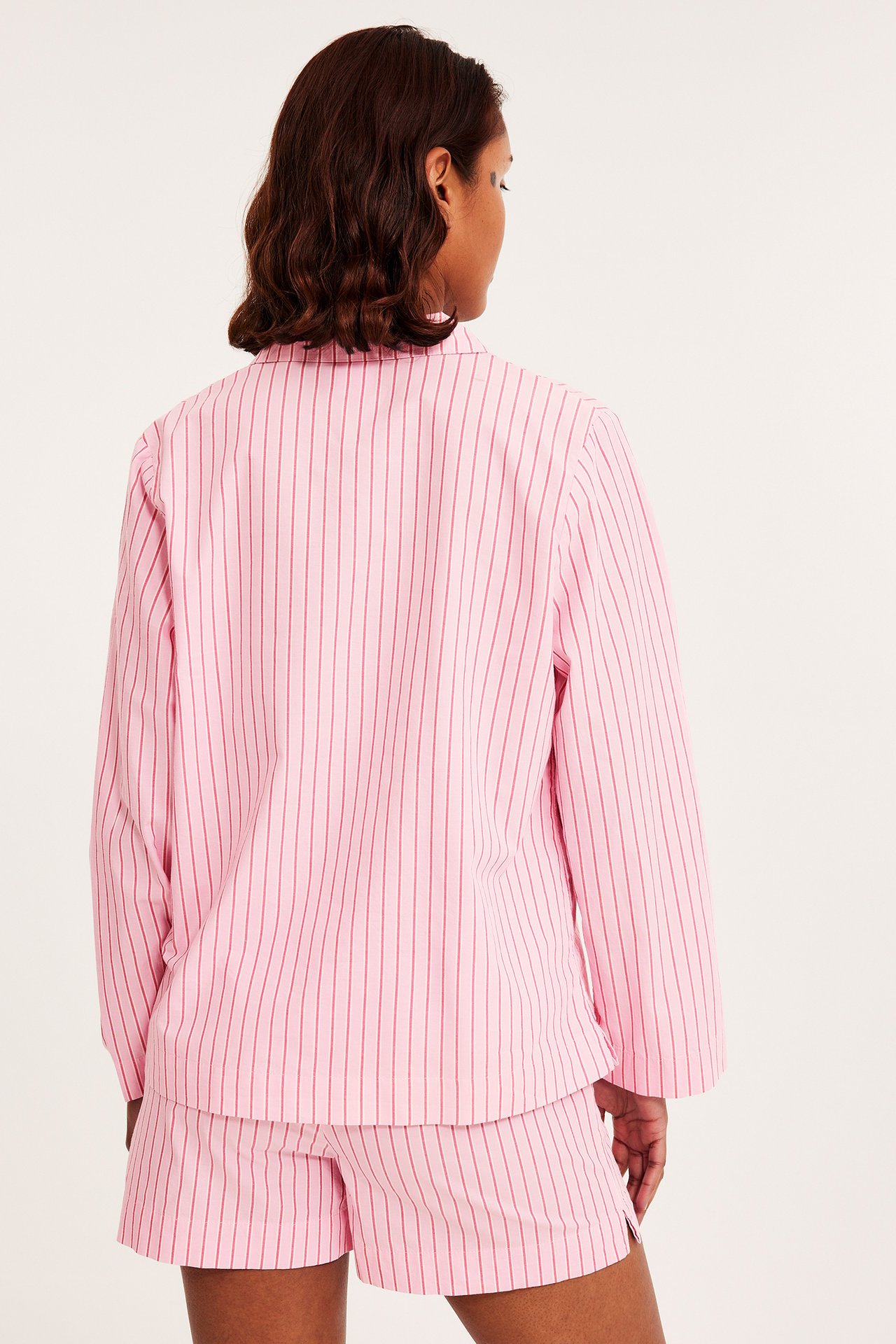 Bakifrån: kvinna i rosa randiga pyjamasshorts i bomull och matchande långärmad pyjamasskjorta.
