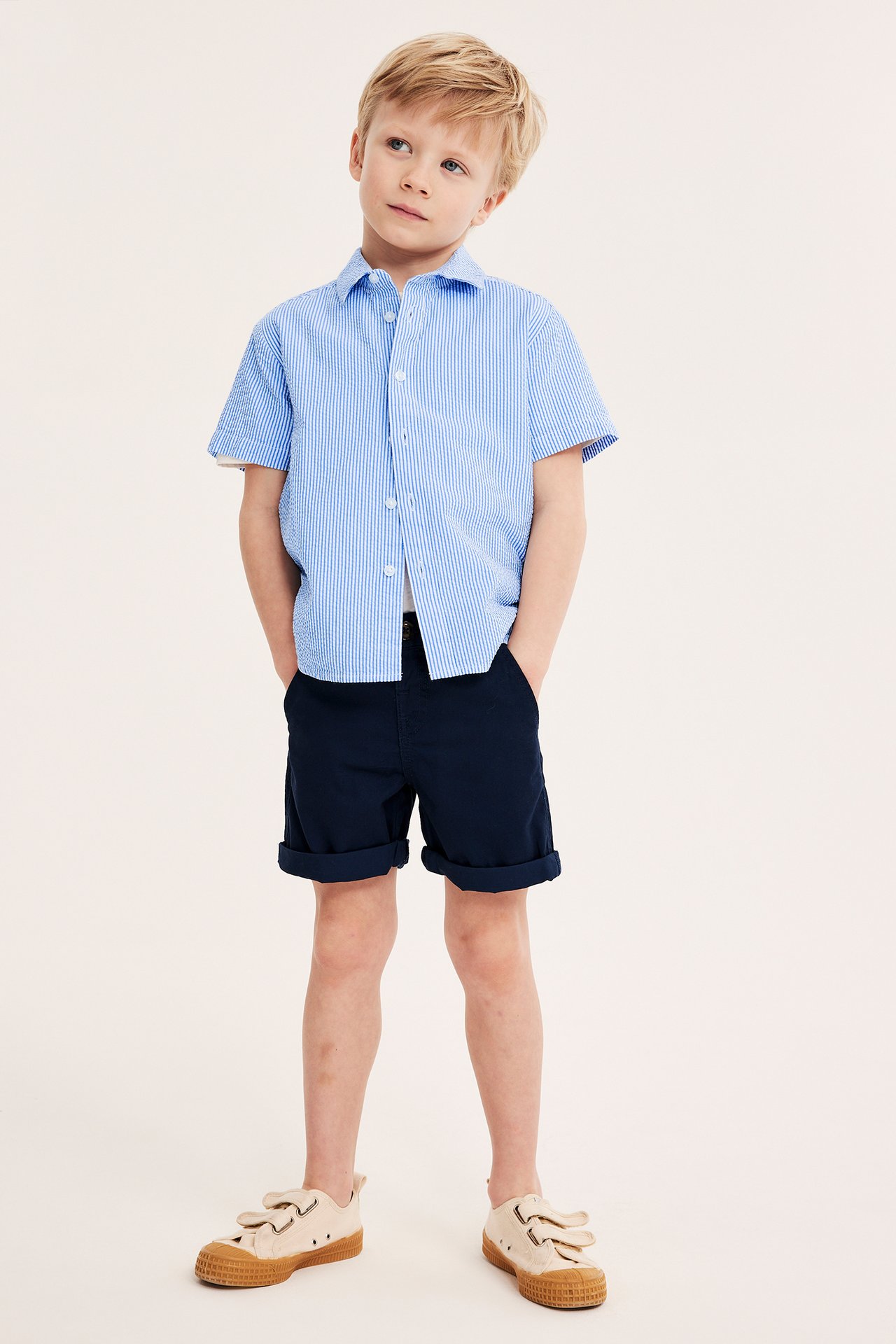 Frontvy av ett barn i en blårandig kortärmad seersuckerskjorta, mörkblå shorts och beiga sneakers.
