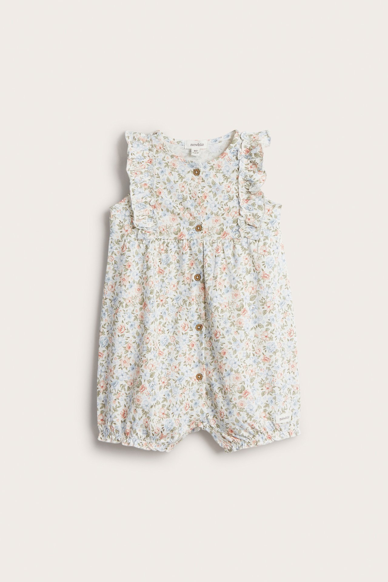 Edestä, luonnonvalkoinen vauvan kukkakuvioinen playsuit, jossa röyhelöt, napit edessä ja nepparit haarassa.