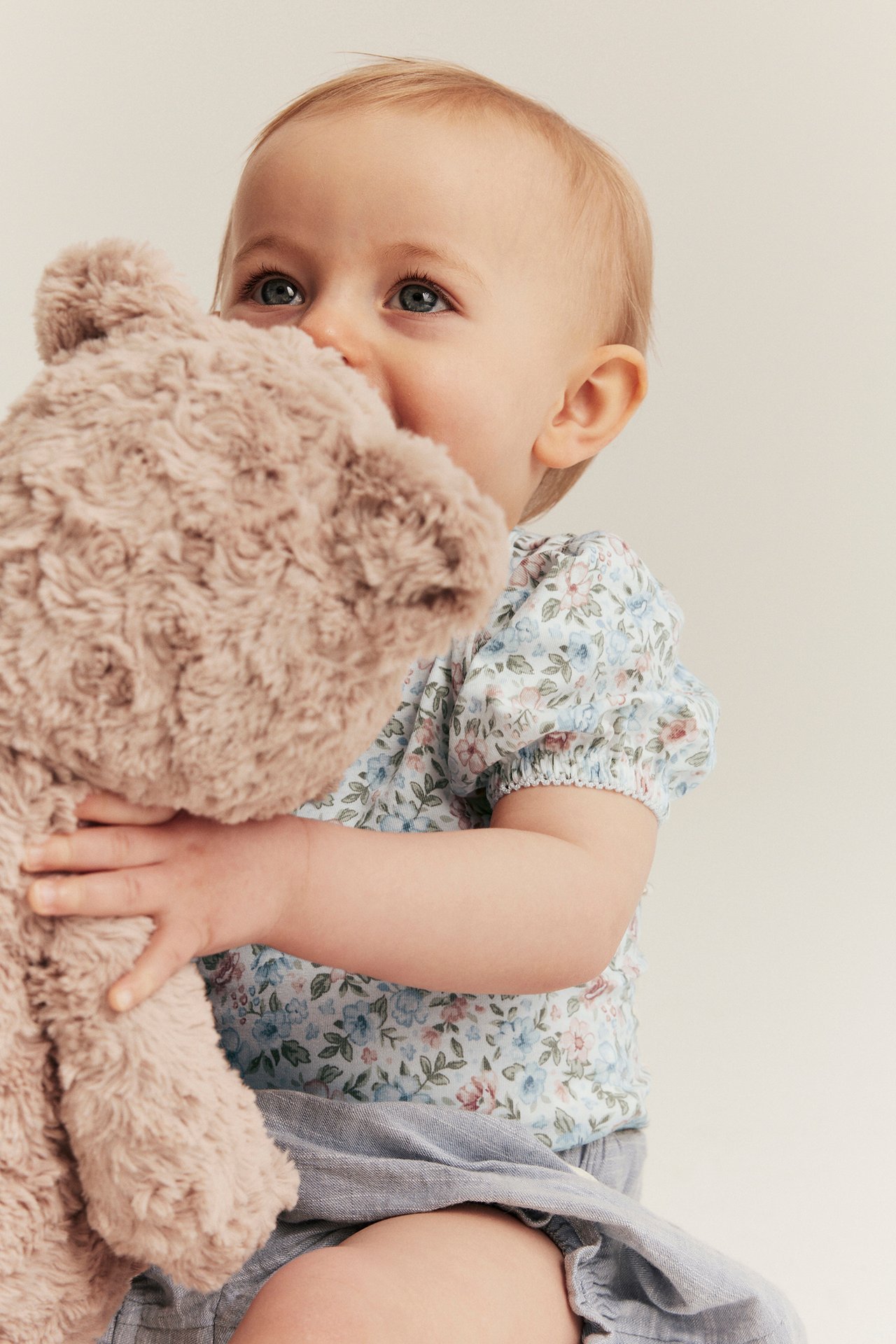 Nærbilde av baby i offwhite blomstret body med korte puffermer, lyseblå shorts, holder en brun teddybjørn.