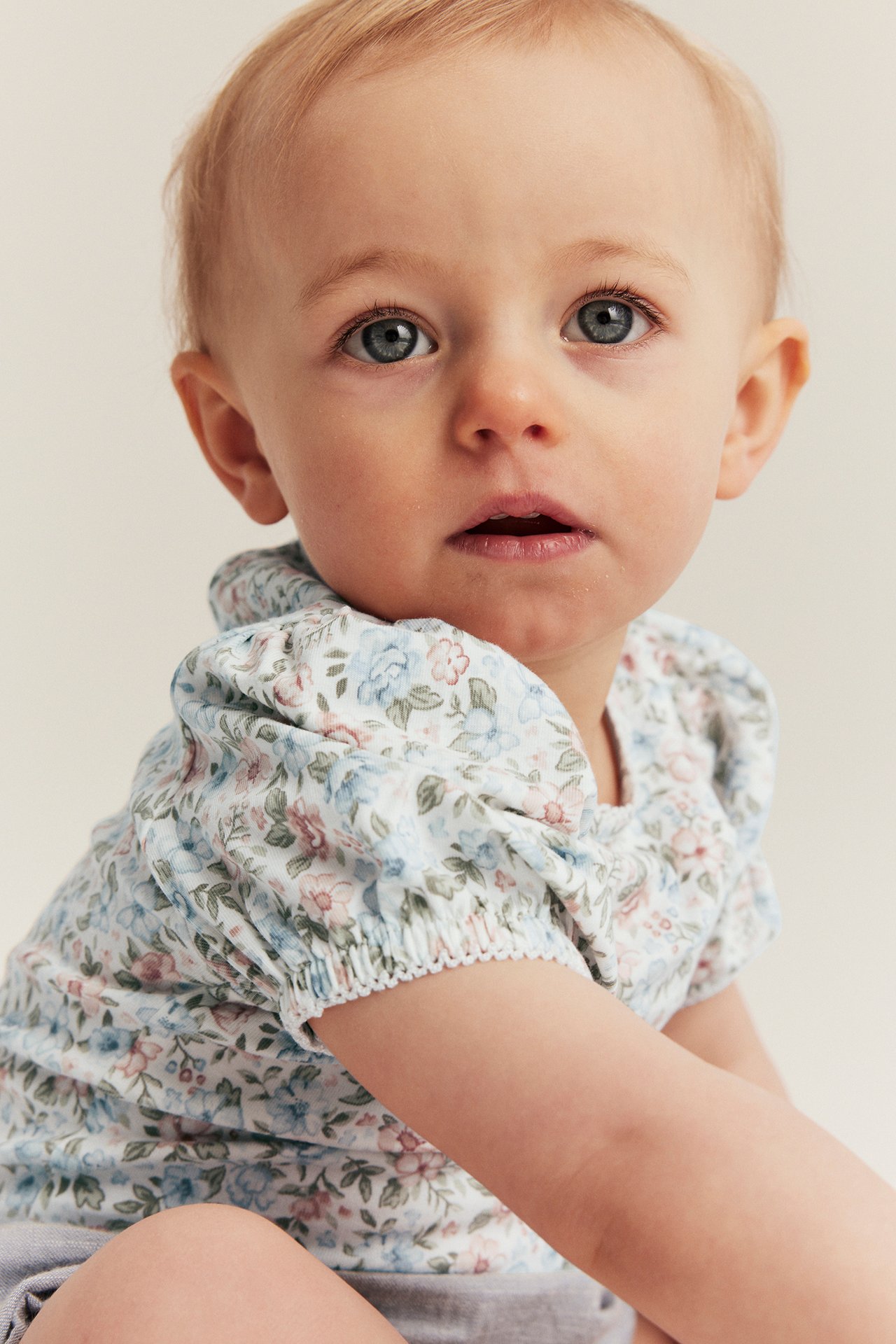 Nærbilde av baby i offwhite blomstret kortermet body med puffermer og strikk i ermekantene.