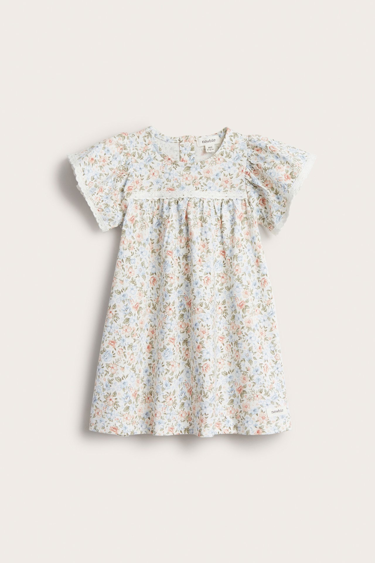 Framsida av en offwhite småblommig trikåklänning för baby med korta ärmar, spetskanter och tryckknappar i ryggen.
