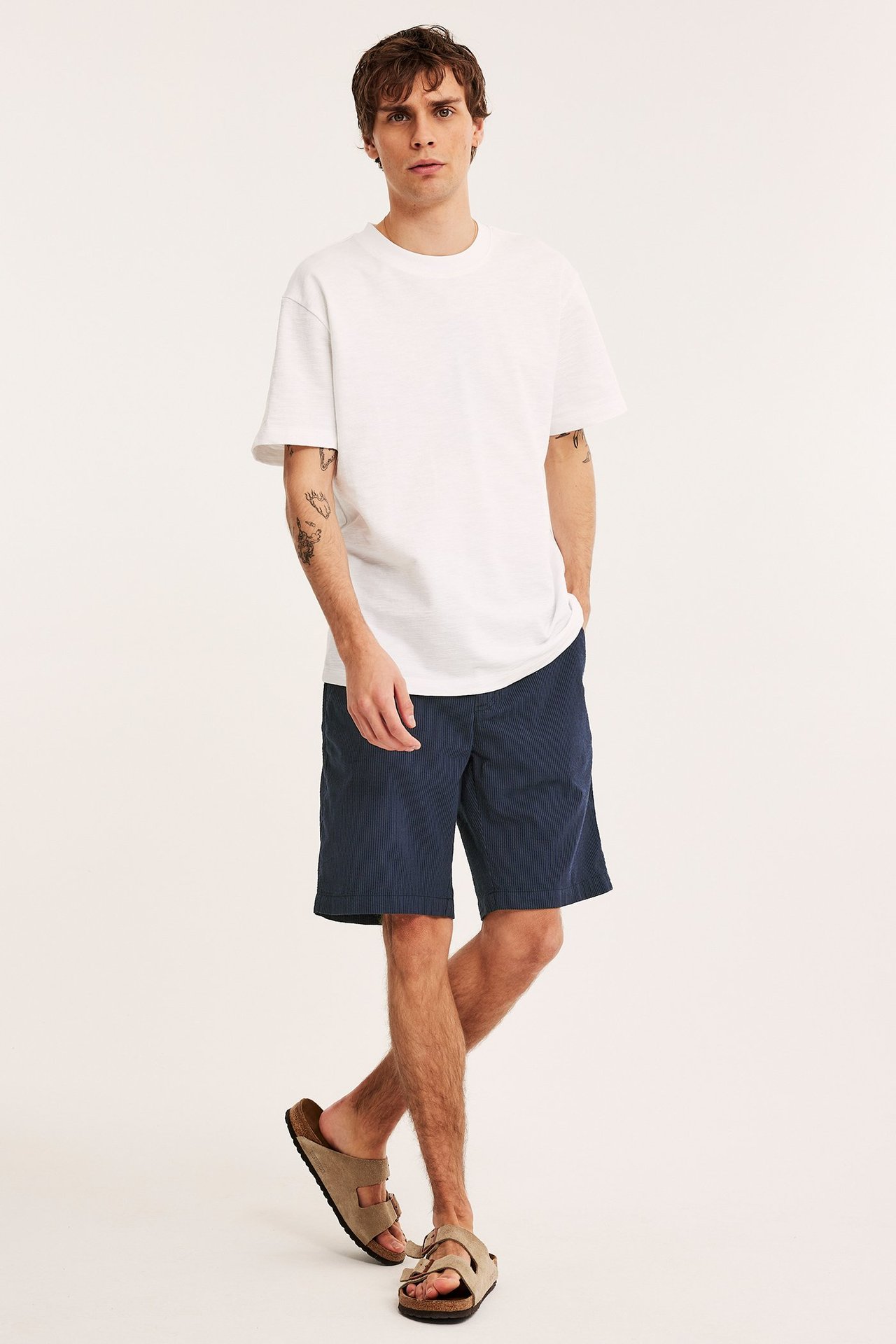 Edestä kuvattu mies yllään tummansiniset raidalliset seersucker-shortsit, valkoinen t-paita ja beiget sandaalit.